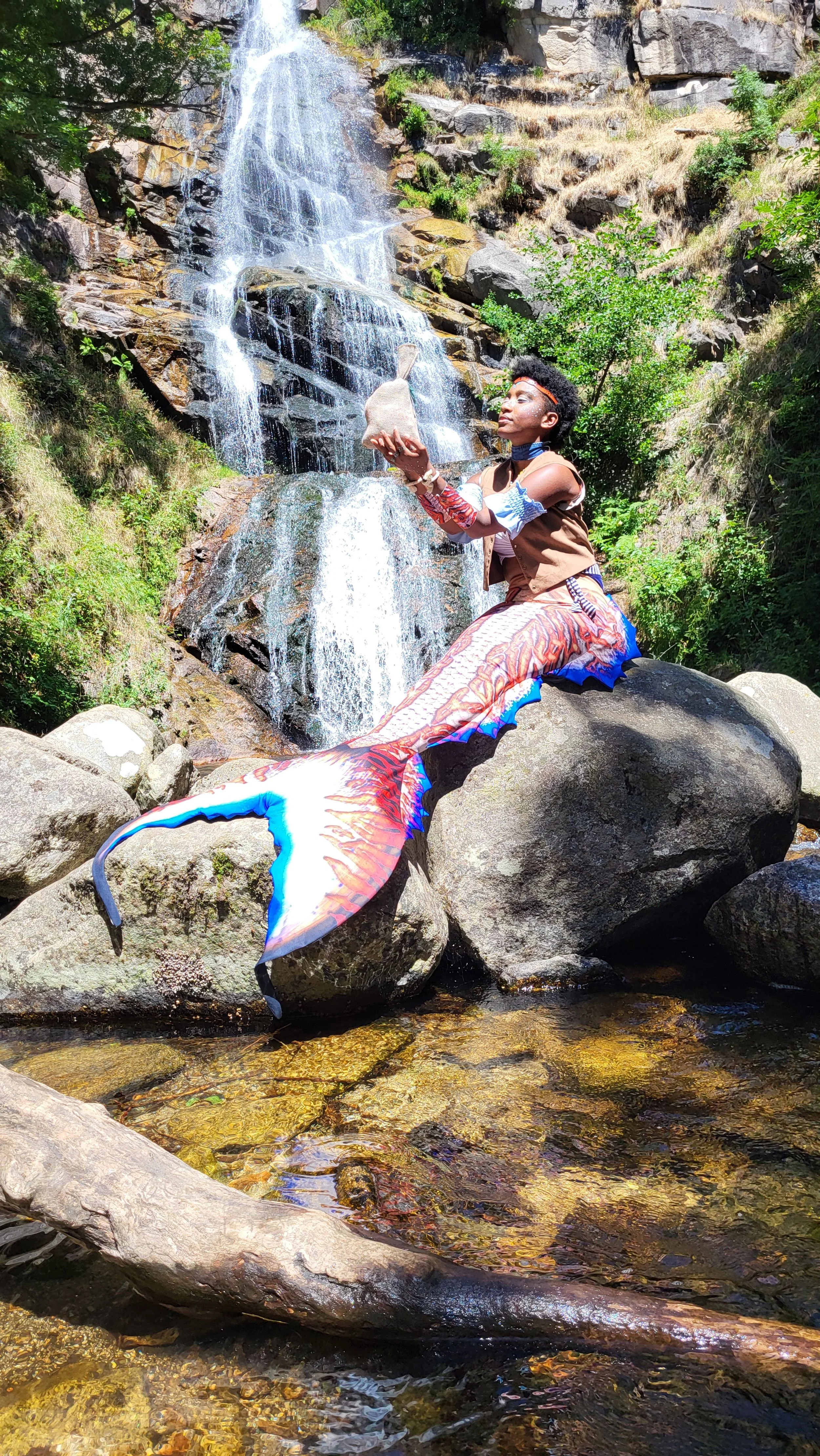 Mermaid Andrina sitting on rocks in a stream with a waterfall in the background. The individual holds a small bag in one hand and has decorative body art and accessories.