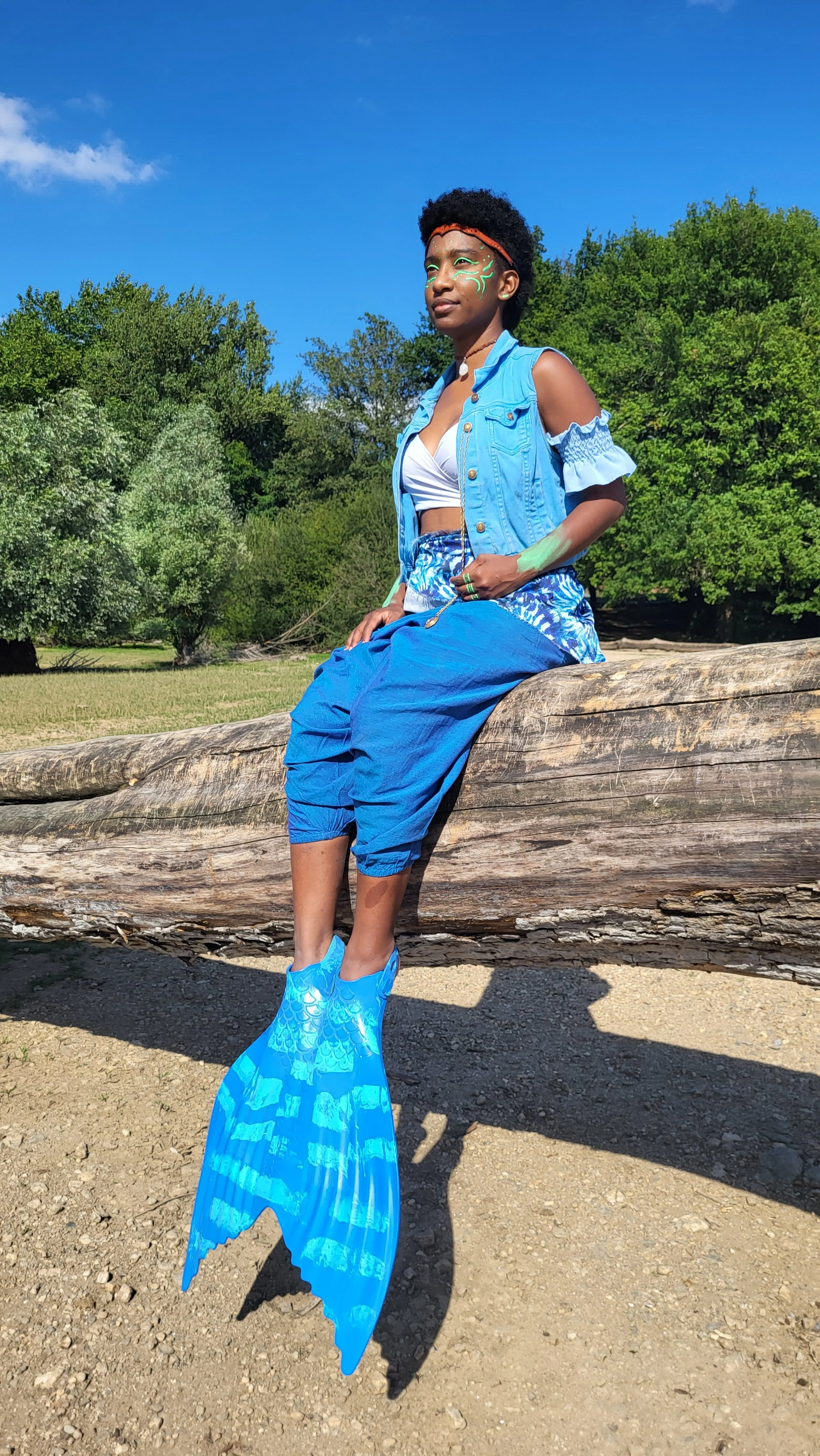 Mermaid Warrior Andrina sitting on a large fallen tree trunk outdoors with greenery and blue sky in the background. She has body art, wears a blue denim vest, a white crop top, and blue pants. She has mermaid fins.