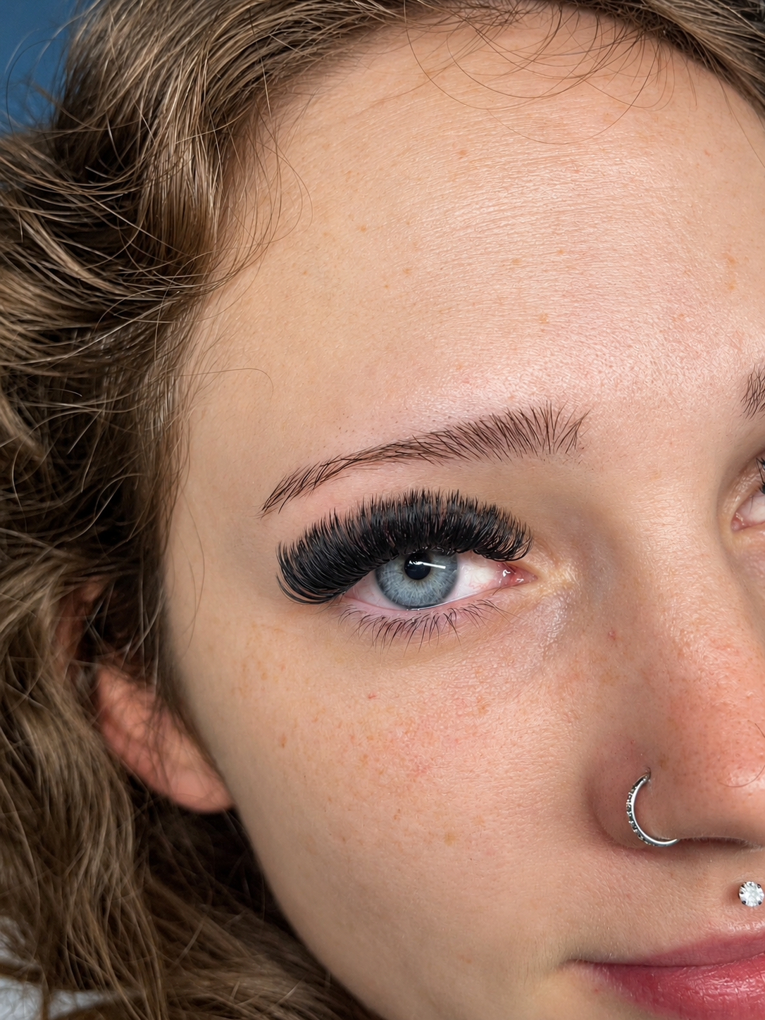 Close-up of a woman's face focusing on her blue eye, with long, thick eyelashes, a small silver nose ring, and her light brown, curly hair.