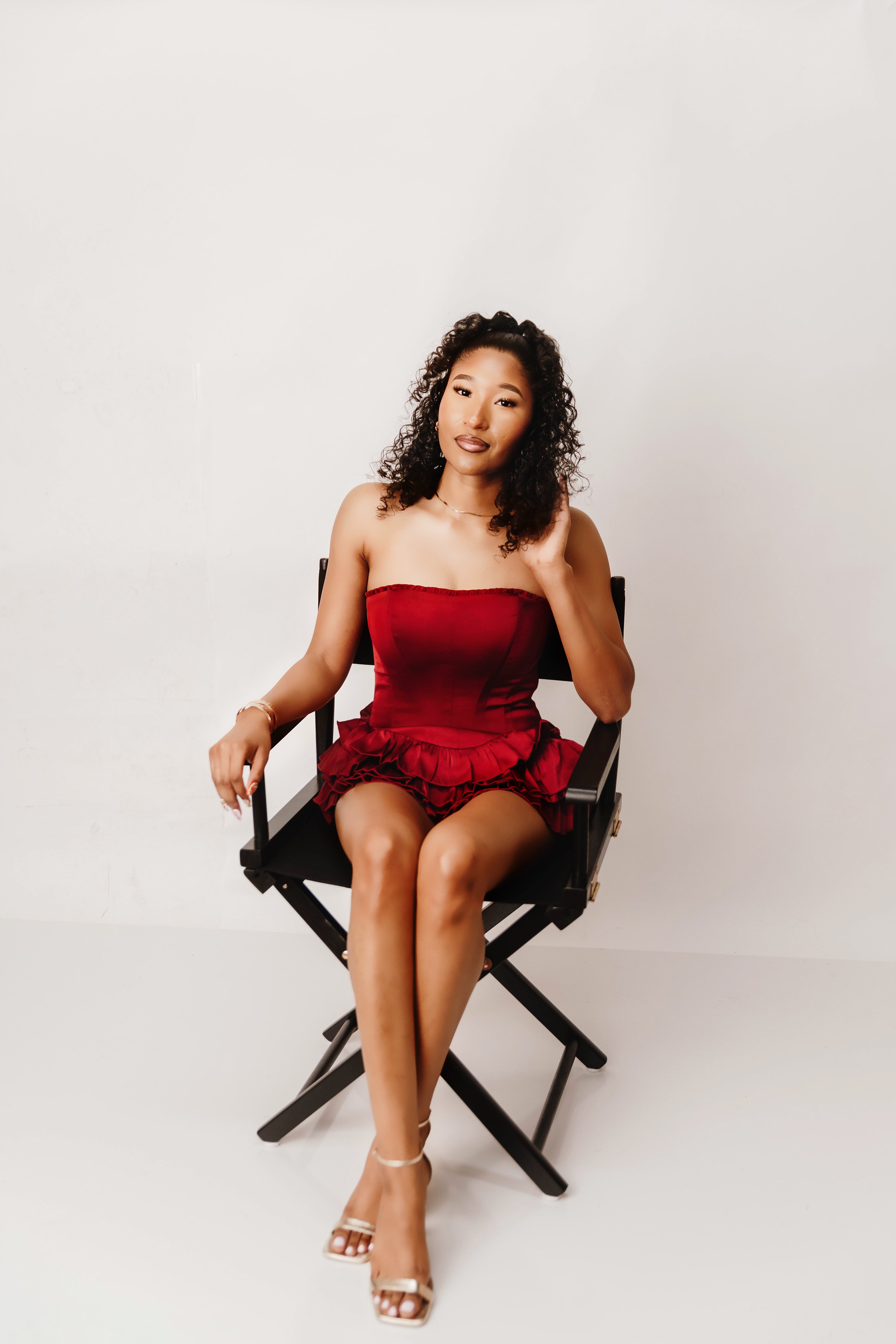 A woman with curly dark hair and light brown skin sitting on a black director's chair with a white background. She is wearing a strapless red dress and beige high-heeled sandals, touching her hair with her right hand and looking at the camera.