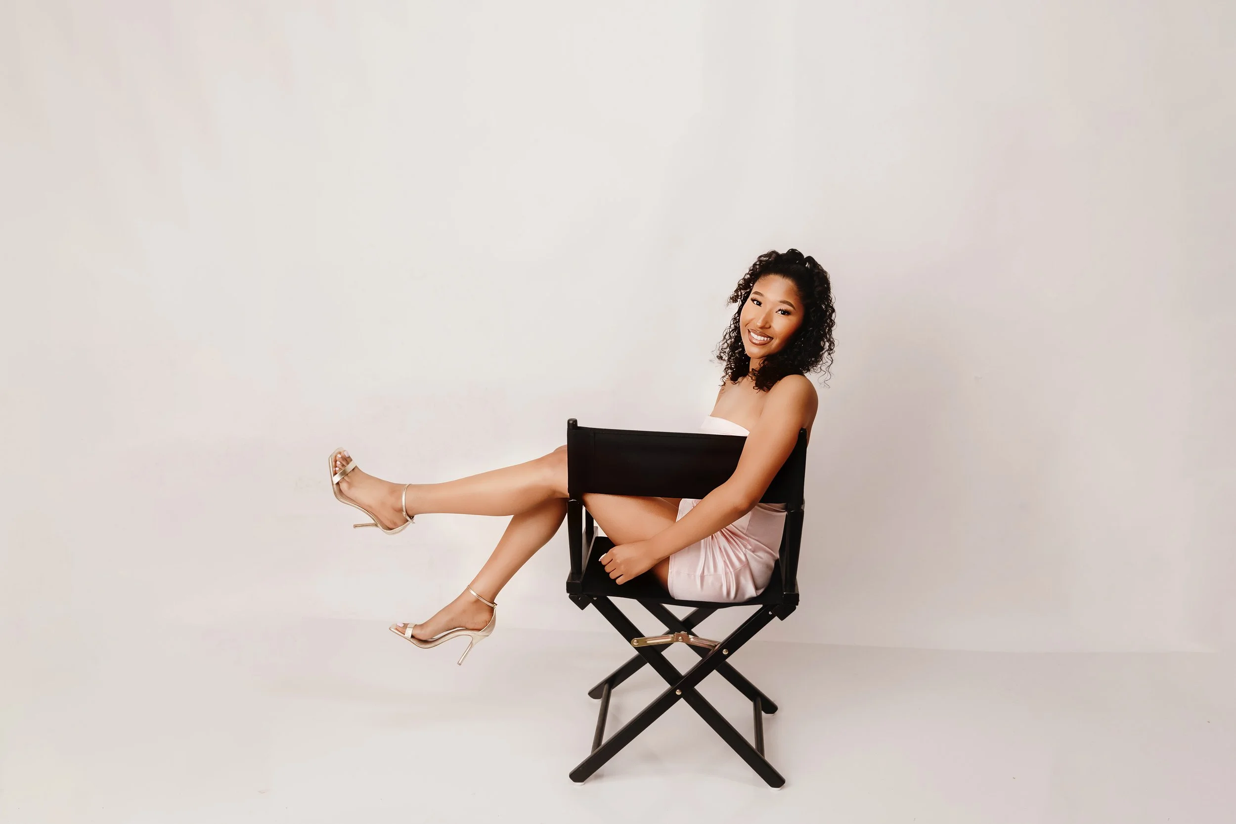 A smiling woman with curly black hair, sitting on a black director's chair, with one leg extended and wearing high heels and a pink dress, against a plain white background.