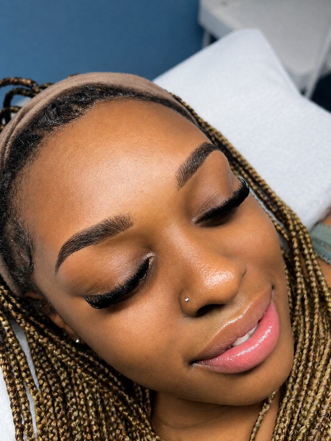 A woman with braided hair, closed eyes, and freshly done eyebrows and eyelashes, lying down with a light pink lip gloss.
