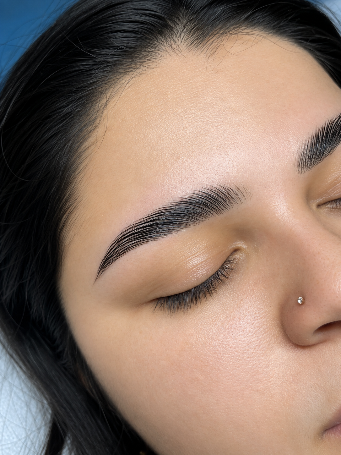 Close-up of a woman's face with closed eyes, showing well-groomed eyebrows, smooth skin, and a nose piercing.