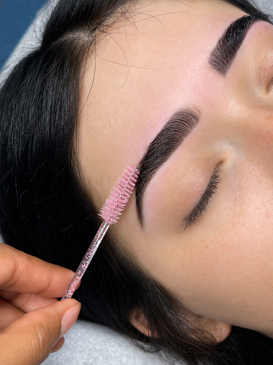 Close-up of a woman receiving eyelash grooming using a pink spoolie brush, with her eyes closed and dark, groomed eyebrows.