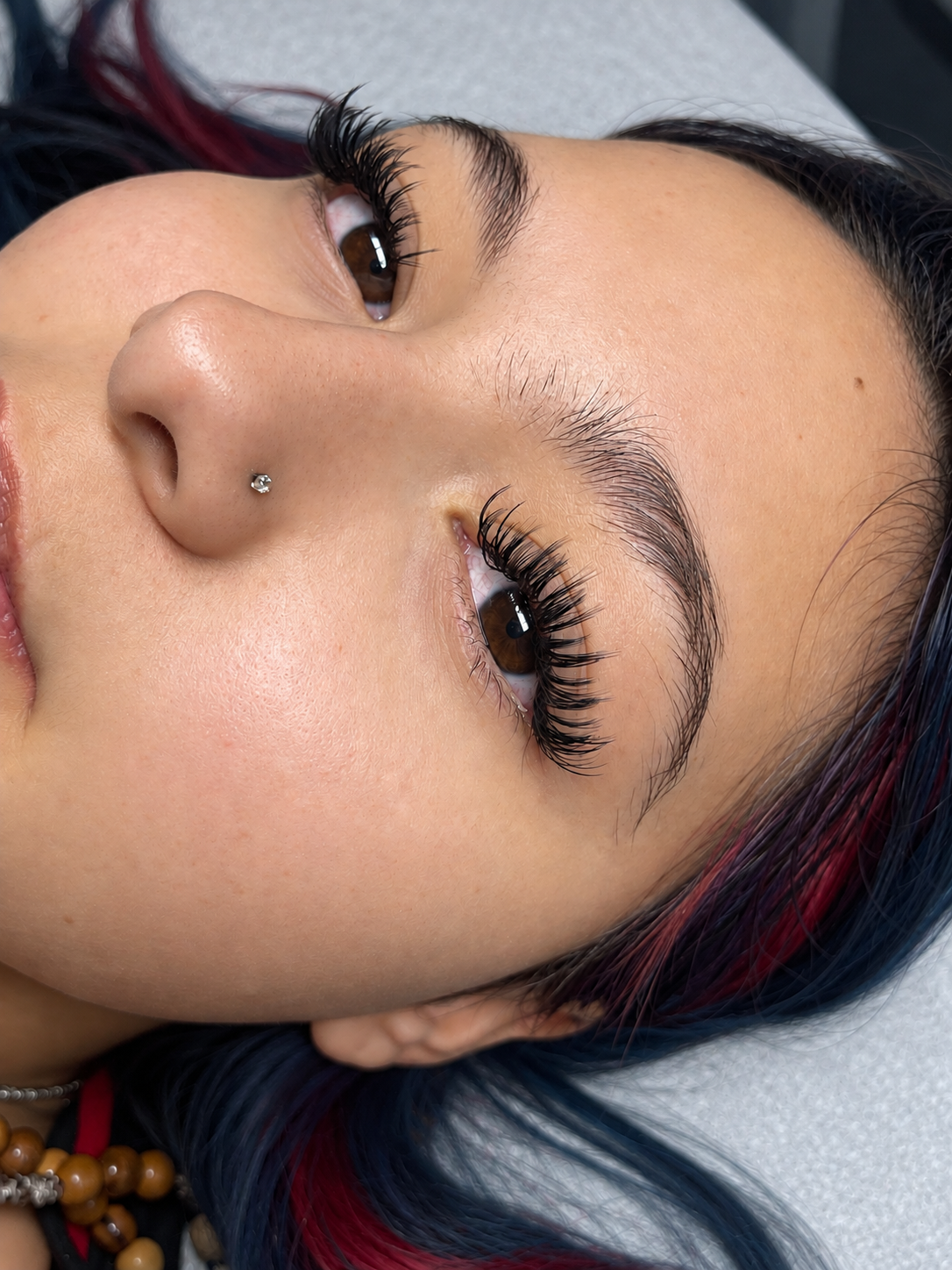 A close-up of a woman's face lying down, showing her brown eyes, long eyelash extensions, nose piercing with a small diamond, and multicolored hair with dark, red, and blue strands.