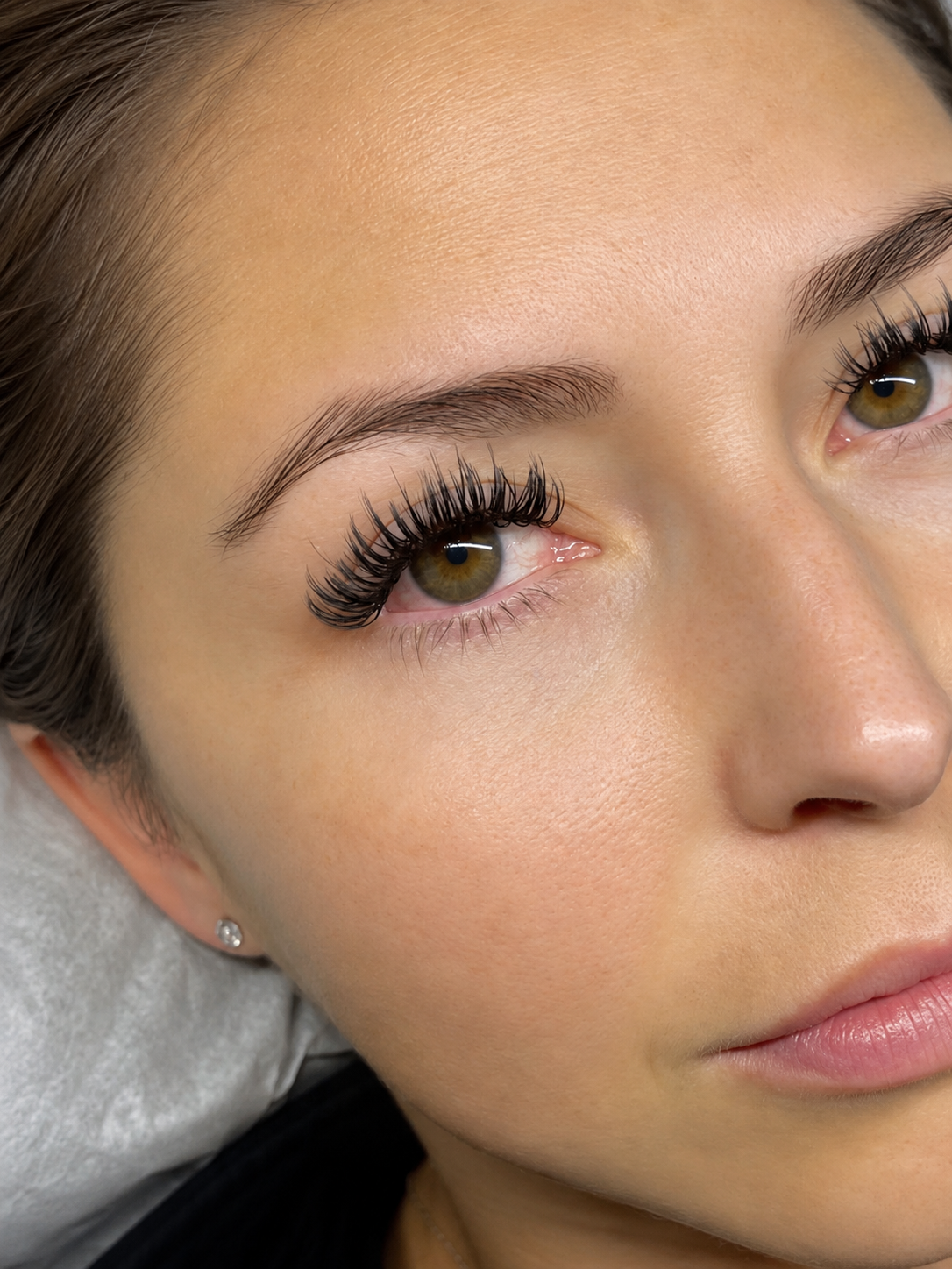 Close-up of a woman's face, showing her brown eyes with long, curled eyelashes, well-groomed eyebrows, clear skin, and a small earring.