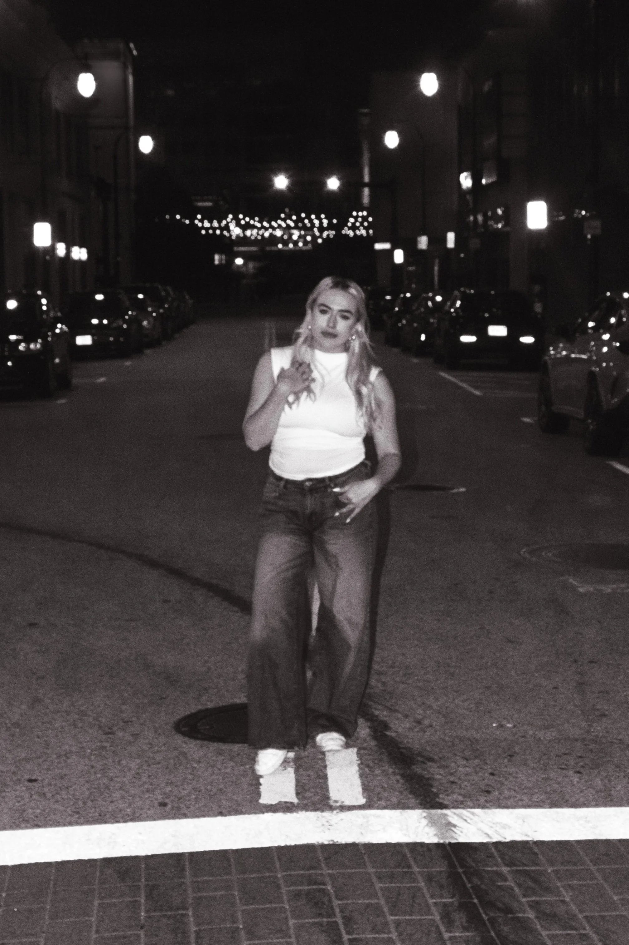 A woman standing on a street at night, illuminated by streetlights, with parked cars lining both sides of the street, wearing a sleeveless top and pants.