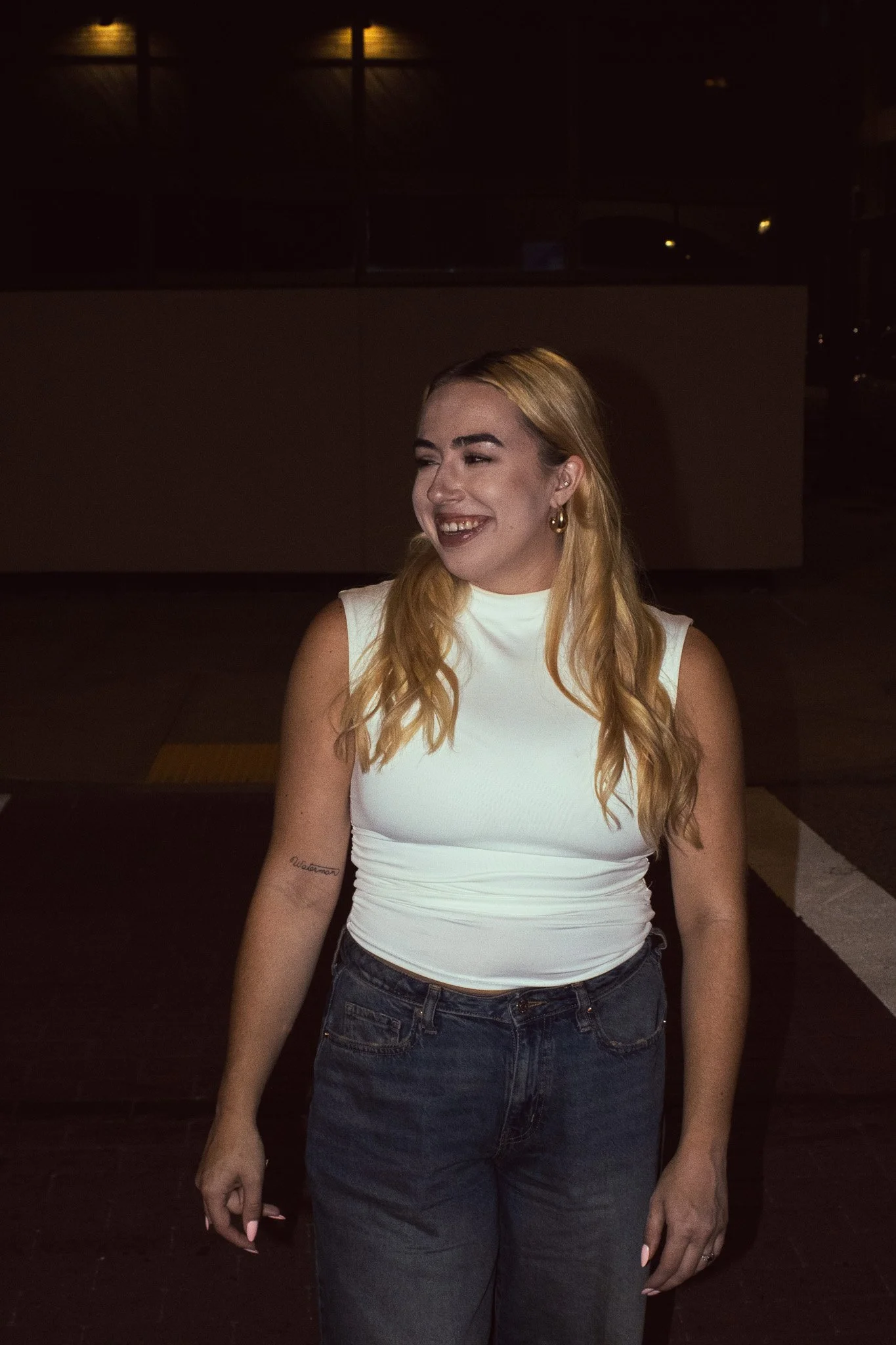 A woman with long blonde hair, smiling and wearing a white sleeveless top and dark jeans, standing in a dimly lit indoor setting.