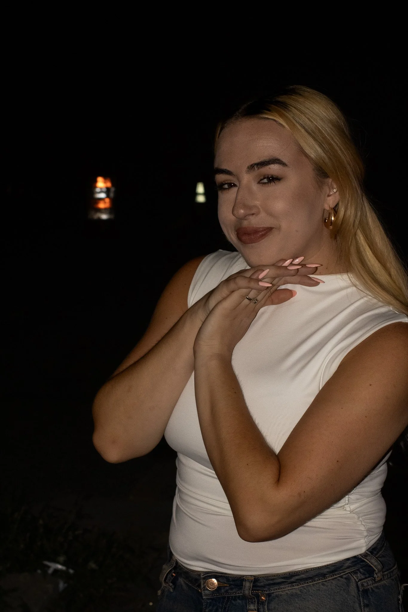 A woman with blonde hair wearing a white top and dark jeans poses with one hand near her chin, smiling slightly against a dark background with a blurred light in the distance.