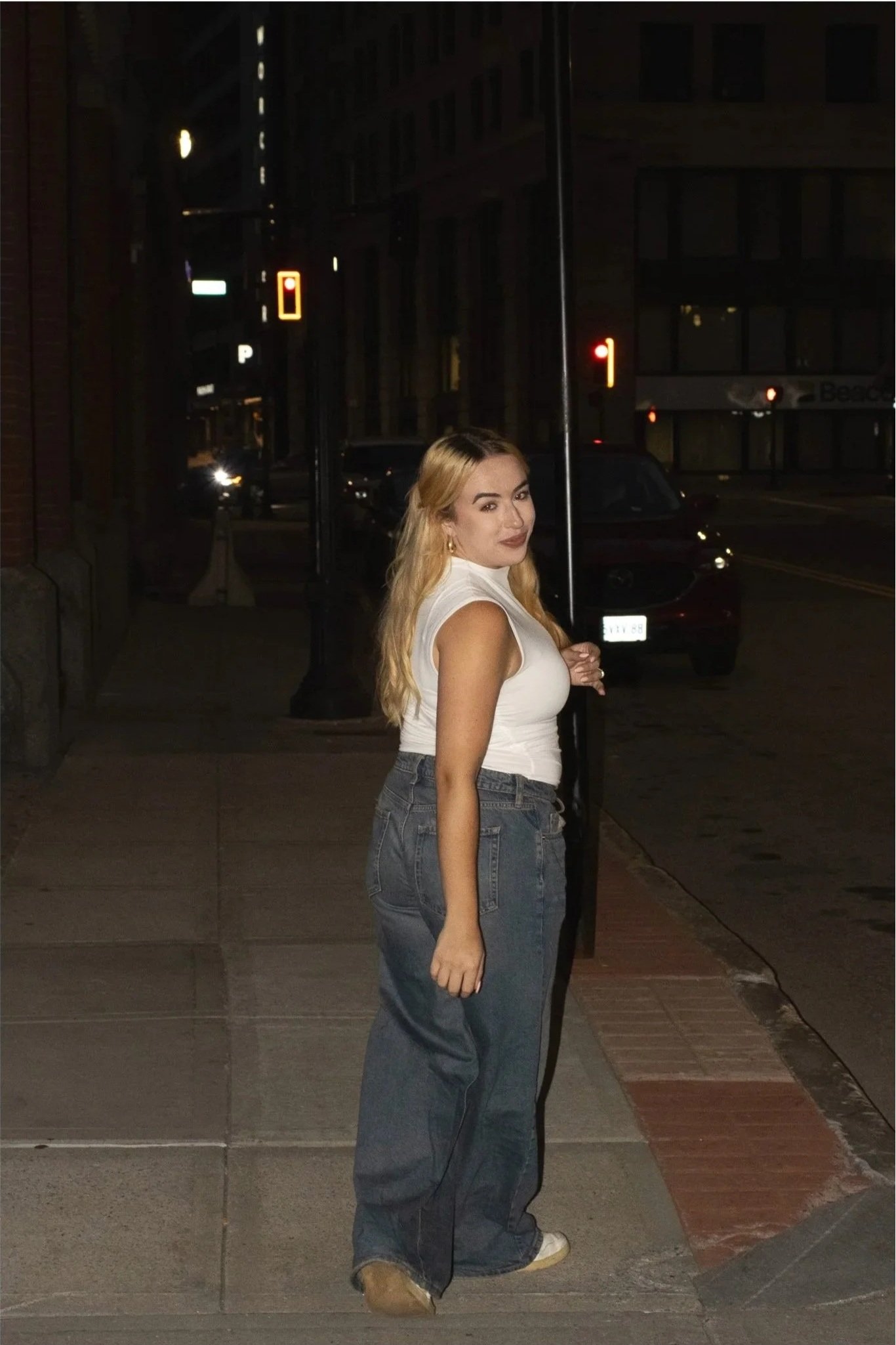 A woman with blonde hair, wearing a white sleeveless top and high-waisted jeans, standing on a city sidewalk at night next to a street pole, with traffic lights and cars in the background.