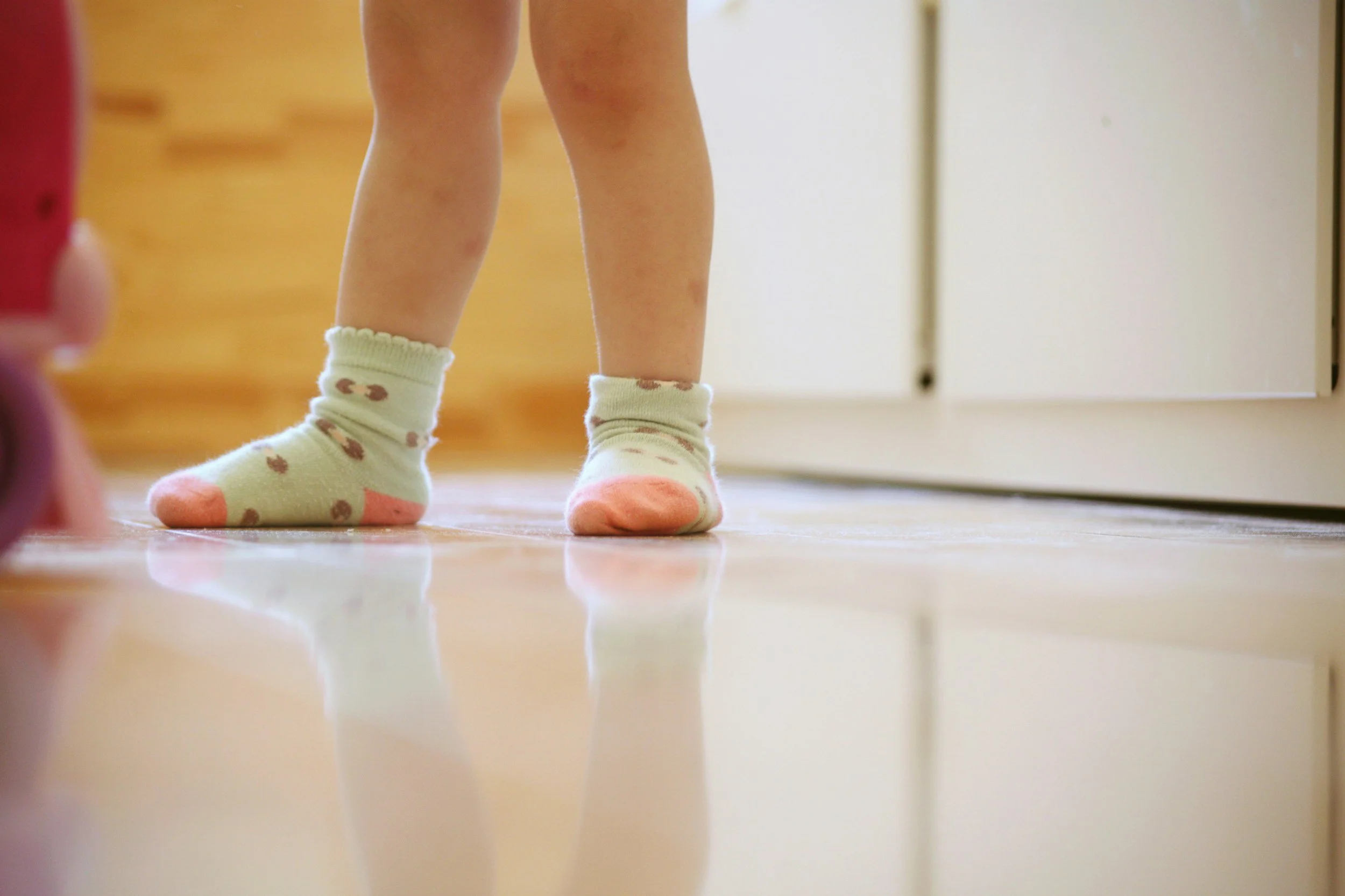 Kind mit weißen Socken mit braunen Flecken steht auf einem Holzboden.