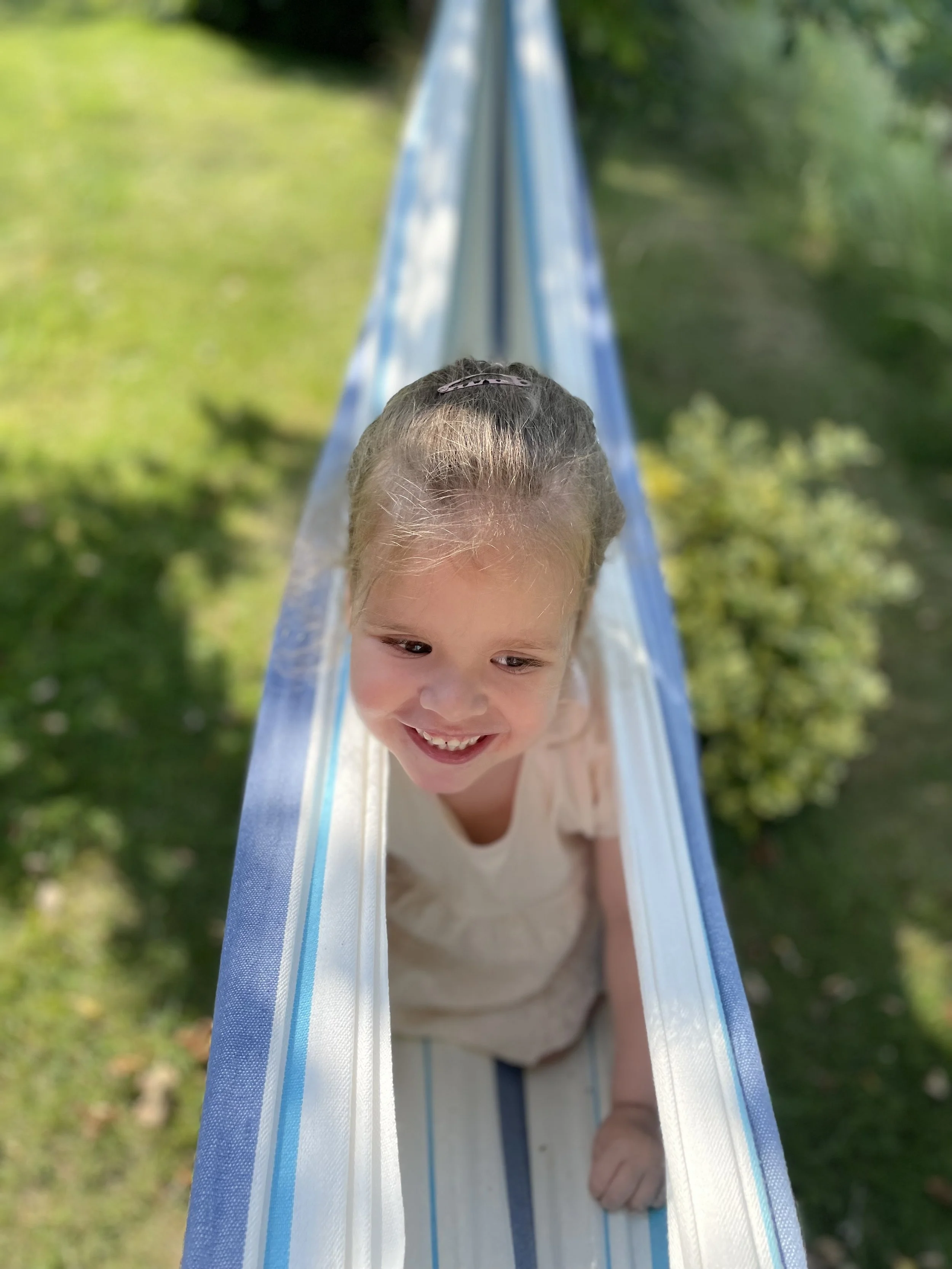 Kind liegt halb in einem weißen und blauen Stofftunnel im Garten, schaut lachend nach vorne, umgeben von Grün.