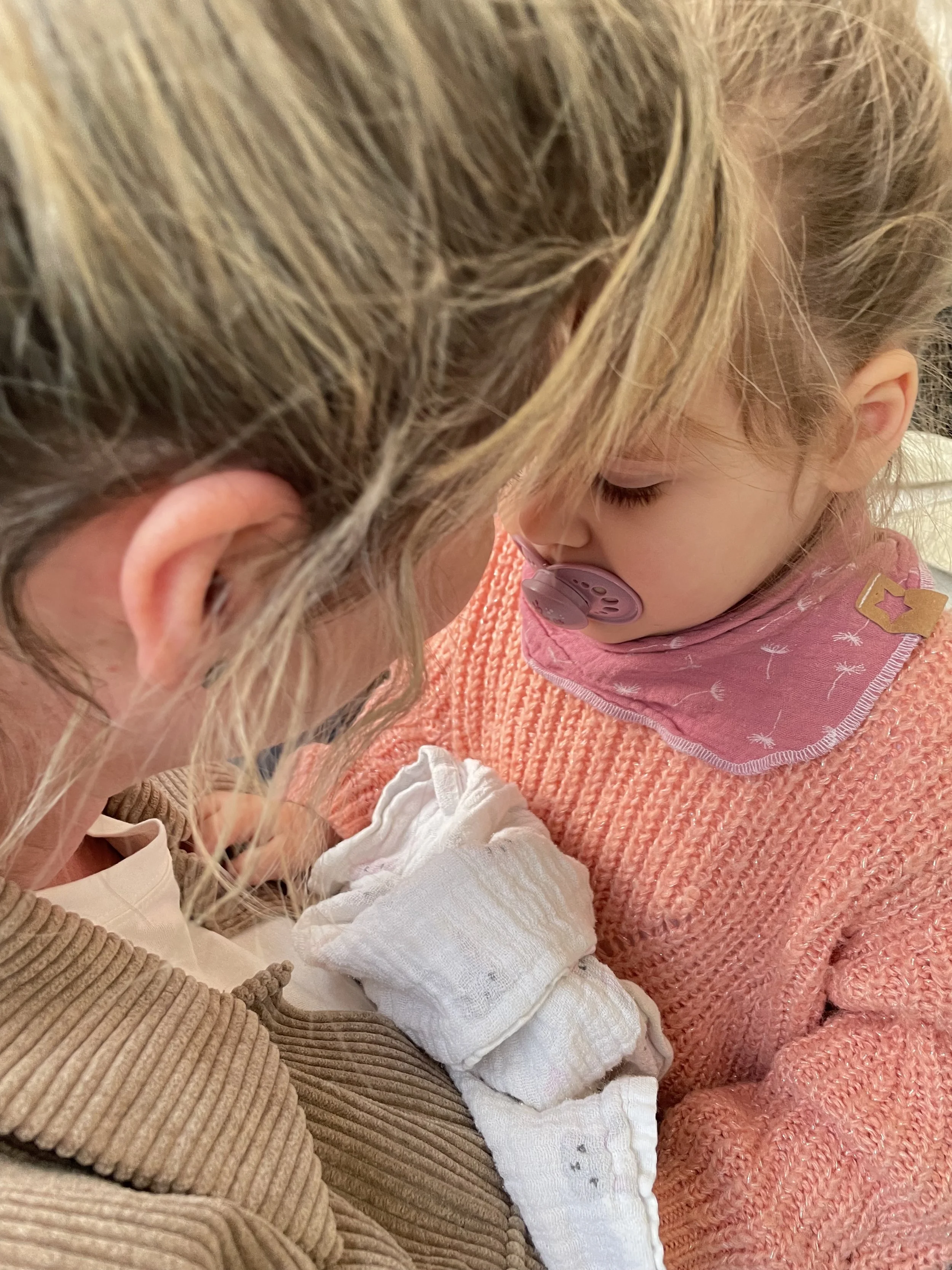 Ein Baby mit Schnuller schläft eng an eine erwachsene Person, vermutlich die Mutter, angekuschelt in einer Nähe bei angenehmer Beleuchtung.