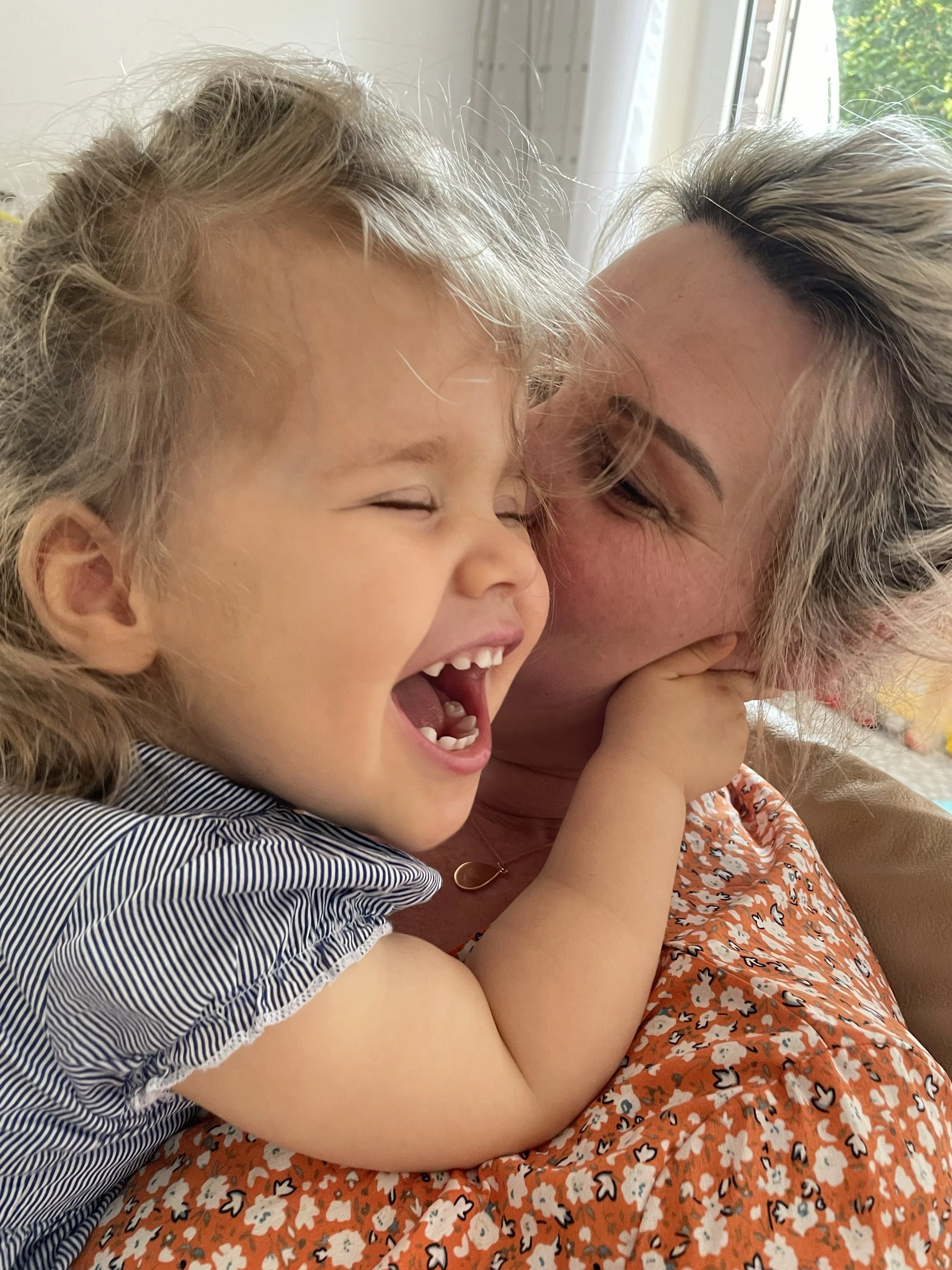 Ein Mädchen und eine Frau lachen eng umeinander, die Frau küsst das Mädchen auf die Wange. Es scheint Freude und Nähe zwischen den beiden zu geben, die vor einem Fenster sitzen.