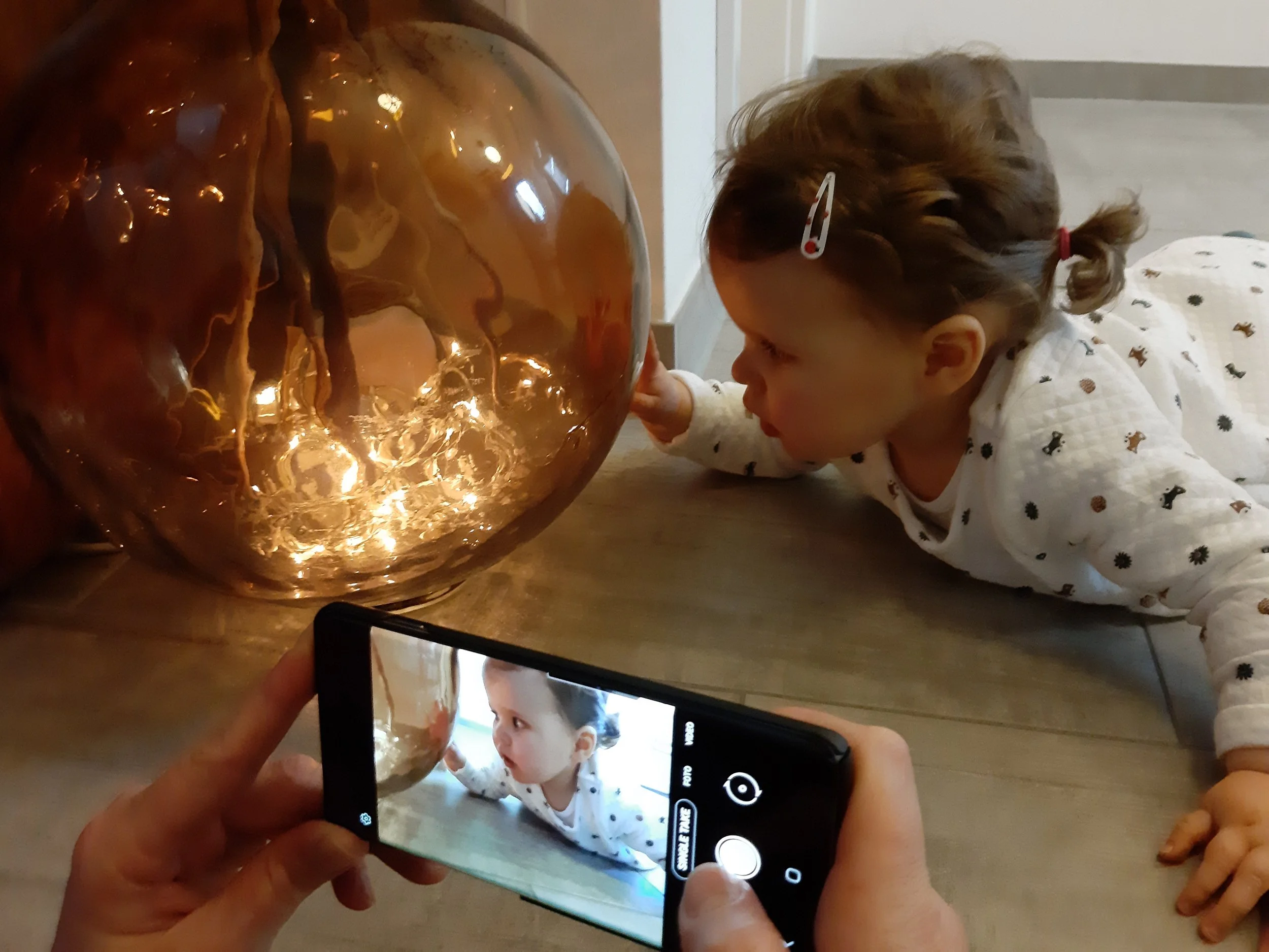 Ein Kind liegt auf dem Boden und schaut auf eine Vase mit goldener Dekoration. Es wird von einer Person mit einem Smartphone fotografiert.