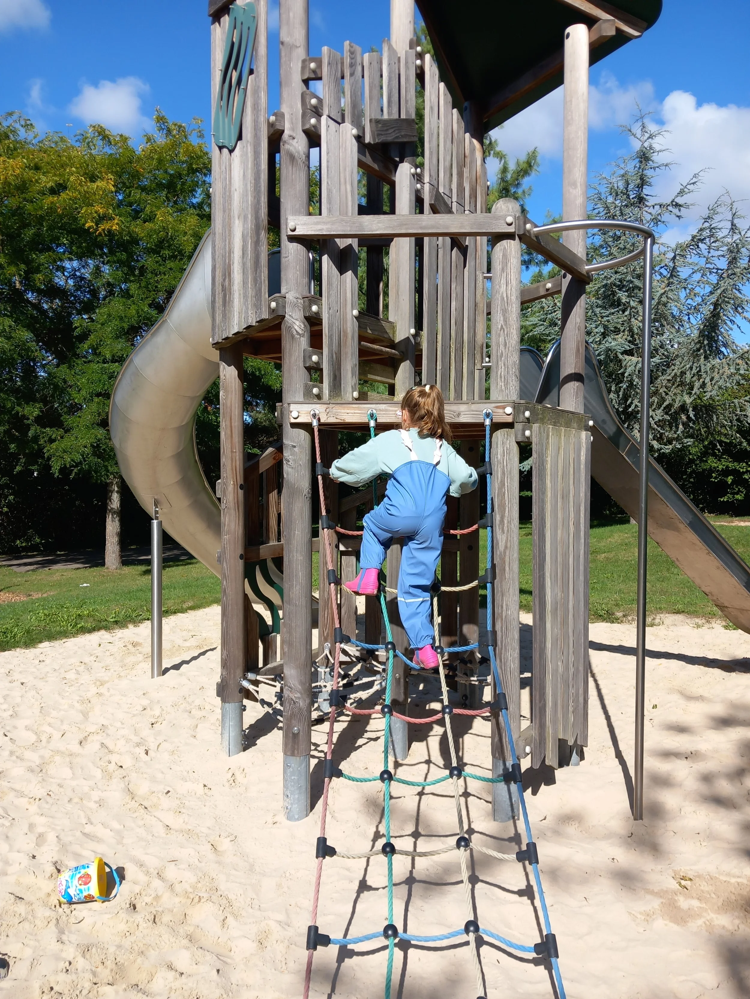 Ein Kind klettert an einer Kletterwand auf einem Spielplatz aus Holz, mit einem Rutsche links und einem Strand im Vordergrund, umgeben von Bäumen und einem blauen Himmel.