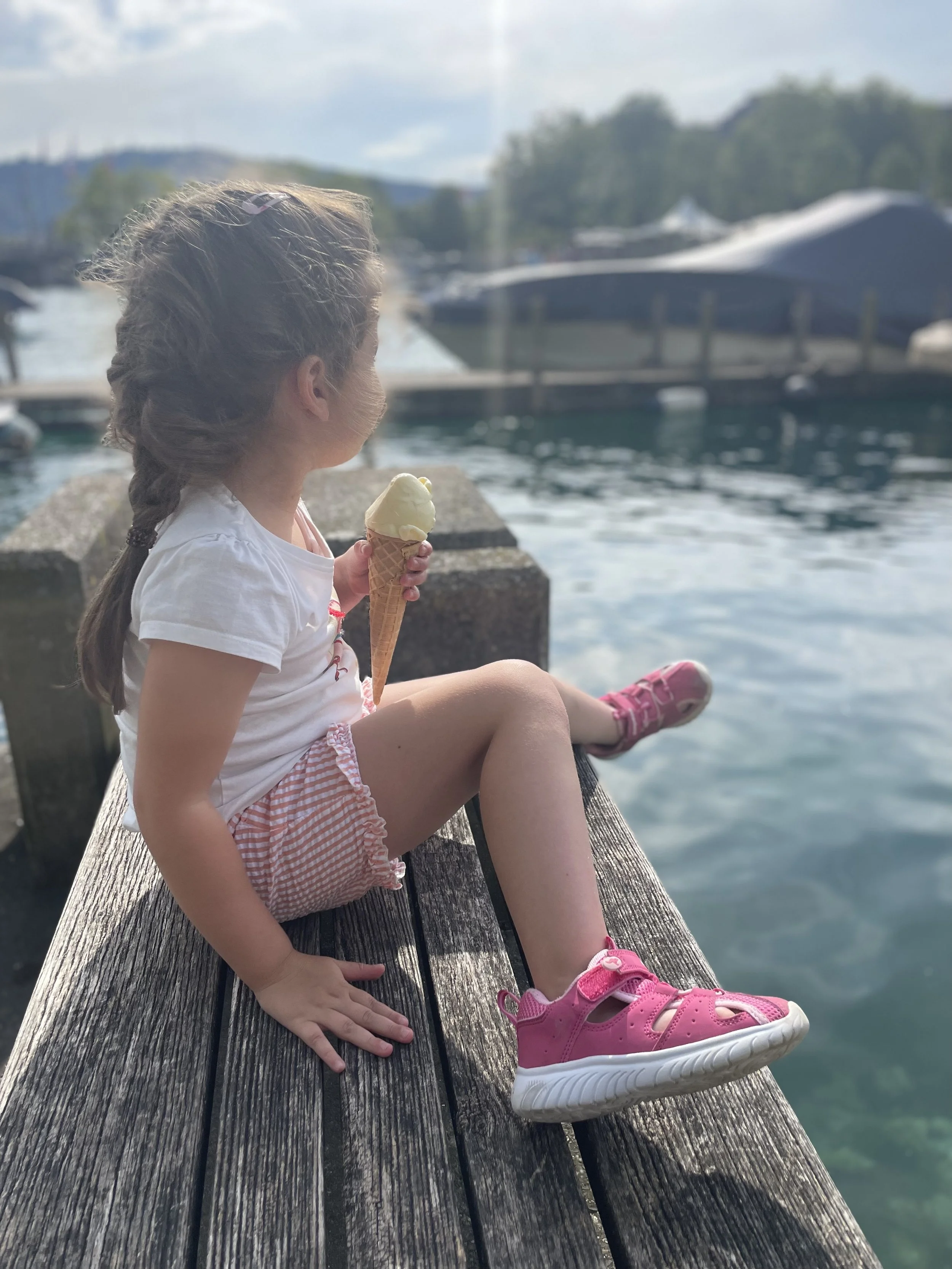 Ein Mädchen mit braunen Haaren, das an einem Steg sitzt und ein Eis essen, blickt auf den See, im Hintergrund Boote und Bäume, an einem bewölkten Tag.