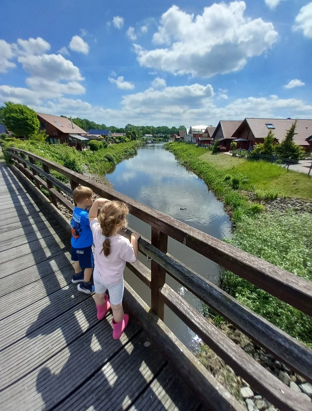 Zwei Kinder schauen von einer Holzbrücke auf einen Fluss in einem Vorort mit Häusern und grüner Vegetation an einem sonnigen Tag.