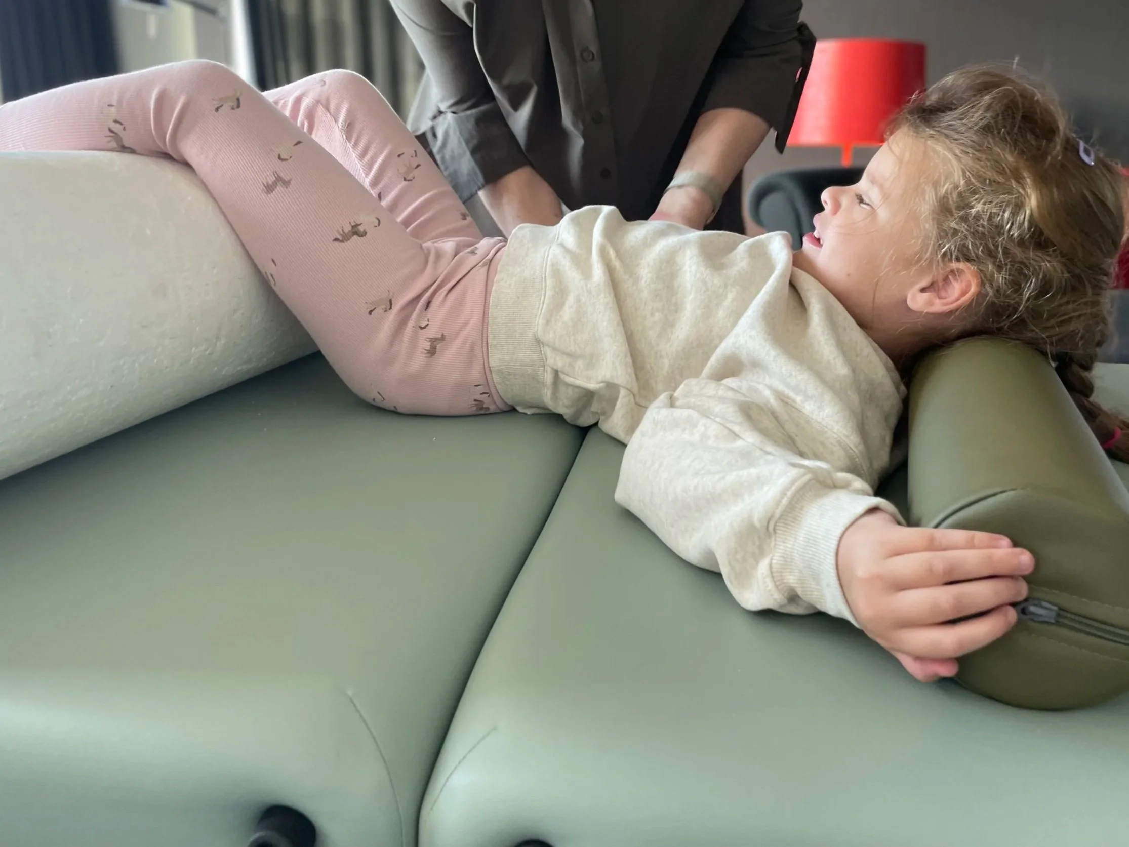 Ein Kind liegt auf einem Untersuchungstisch, während eine Person neben ihm steht und eine Untersuchung durchführt. Das Kind trägt eine helle Jacke und pinke Hosen mit Mustern, das Gesicht ist lachend. Das Umfeld scheint eine medizinische Einrichtung zu sein.