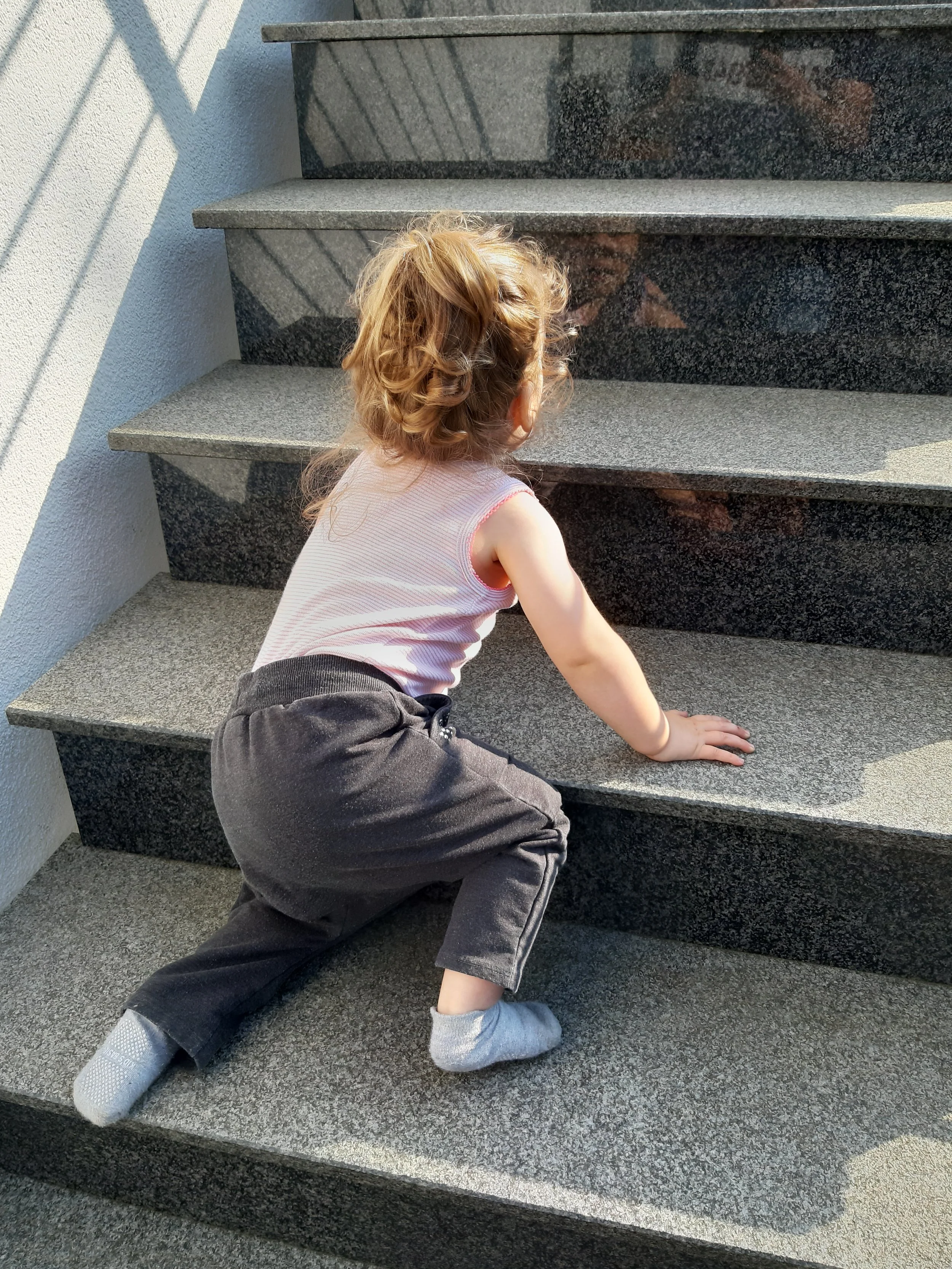 Ein junges Kind mit lockigem braunem Haar, in einem weißen Tanktop und schwarzen Hosen, kriecht auf grauen Stein-Treppen im Freien, Sonnenlicht und Schatten sichtbar.