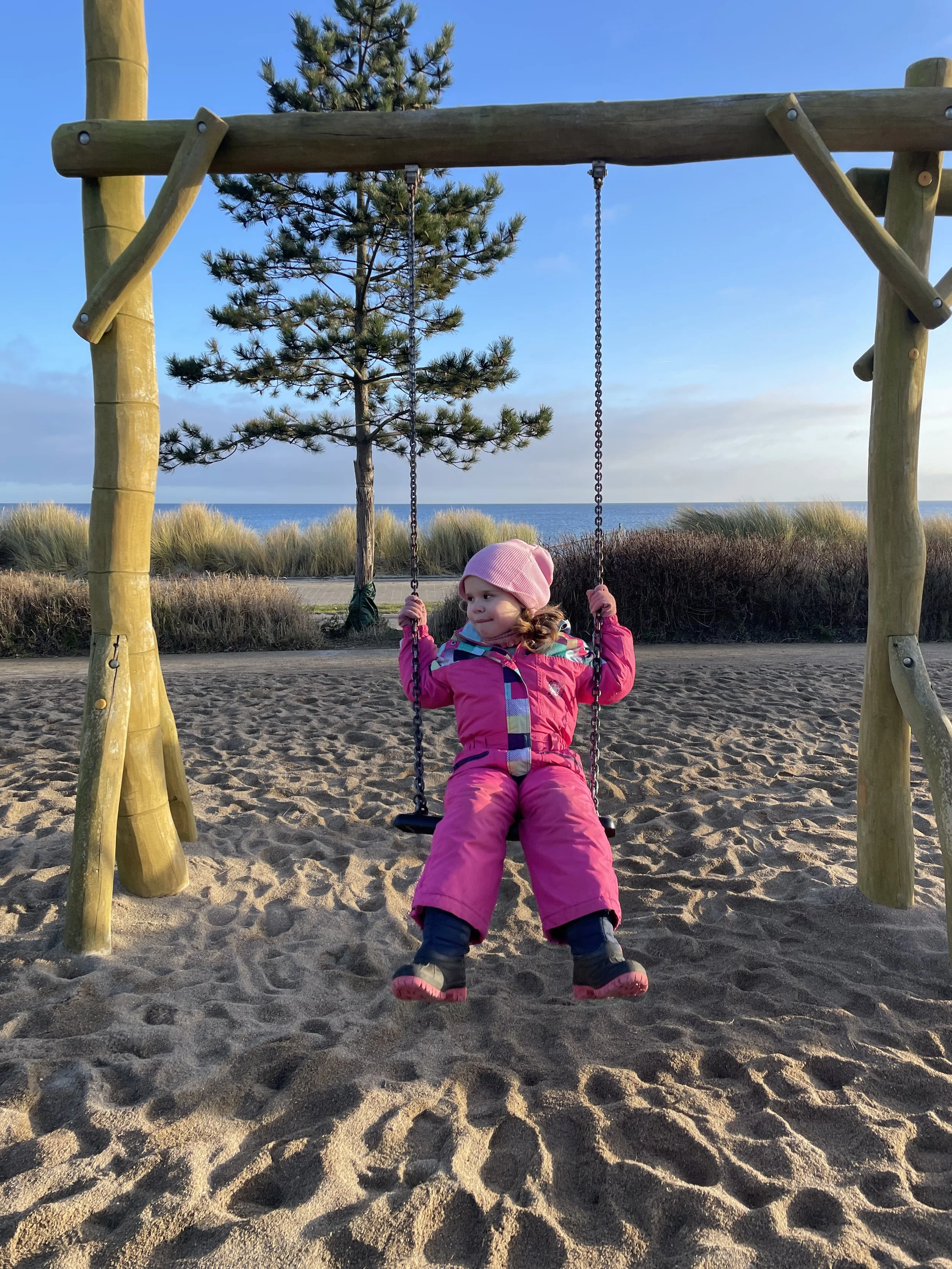 Ein junges Mädchen in einer pinkfarbenen Winterjacke und Mütze sitzt auf einer Schaukel im Sand, im Hintergrund ein Baum, Gras und die Nordsee bei Sonnenuntergang.
