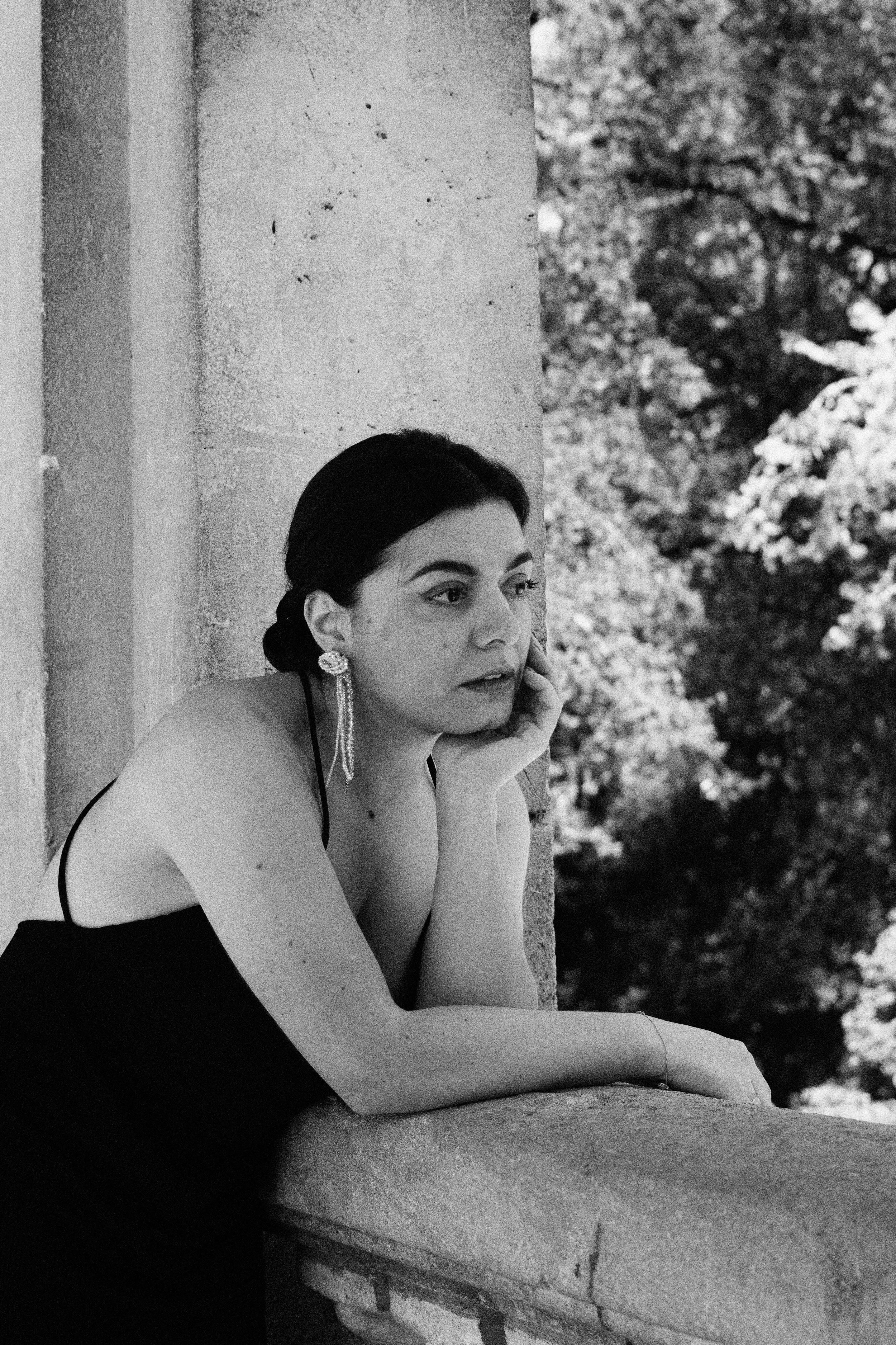 Une femme avec des boucles d'oreilles pendantes, portant une robe noire, est assise contre un mur en pierre, avec des arbres en arrière-plan, en noir et blanc.