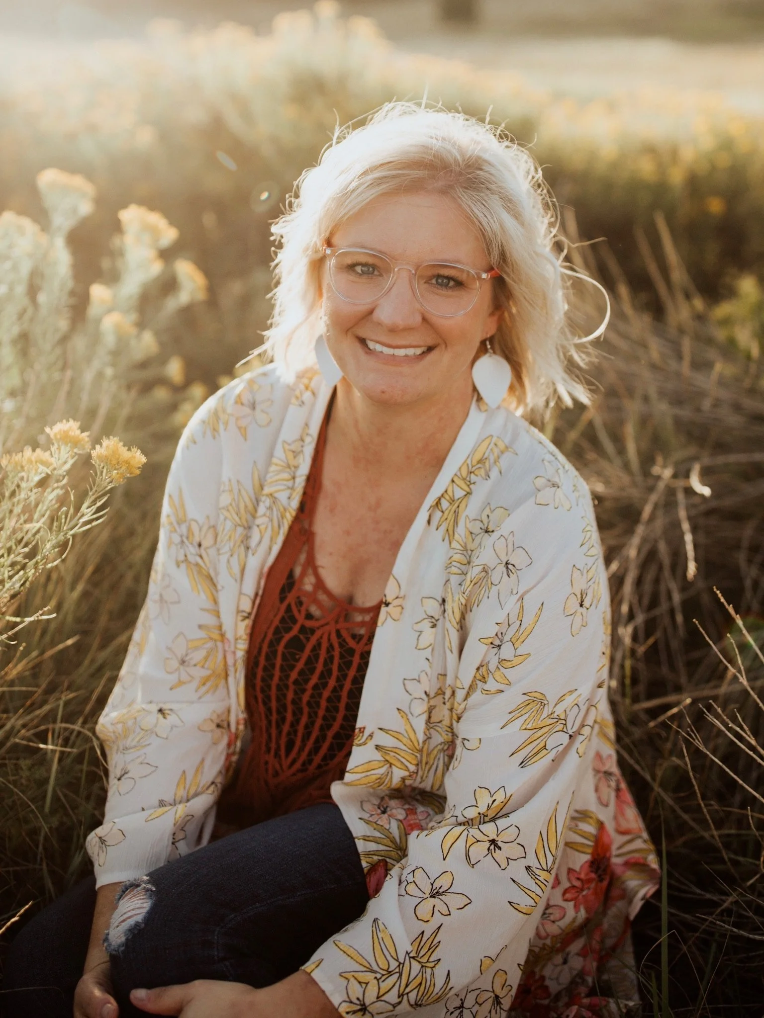 A smiling woman with blonde hair and glasses sitting outdoors among tall grasses and wildflowers, wearing a floral-patterned white kimono-style jacket and a red top.