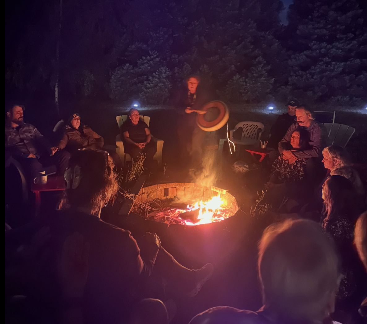 A circle of community members gathered around a fire at night, one raising a hand drum.