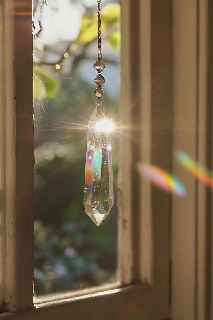 A sunlit hanging crystal prism near a wooden window, with sunlight creating rainbow reflections on the glass and surrounding area.