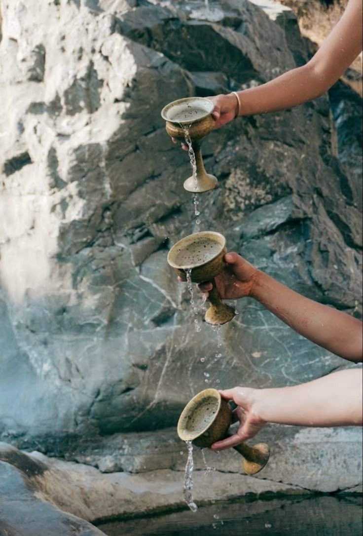 Three hands pouring water from cup to cup down a rocky slope.