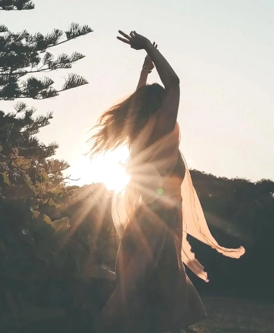 A woman dancing outdoors at sunset, with her arms raised above her head, backlit by the sun with rays shining through her figure, surrounded by trees and foliage.