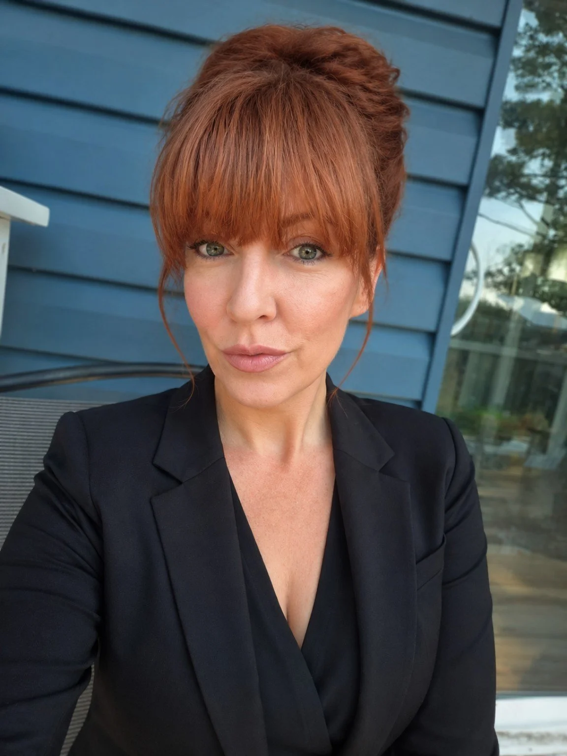A woman with red hair styled in an updo with bangs, wearing a black blazer, taking a selfie outside near a blue house with siding and large windows.
