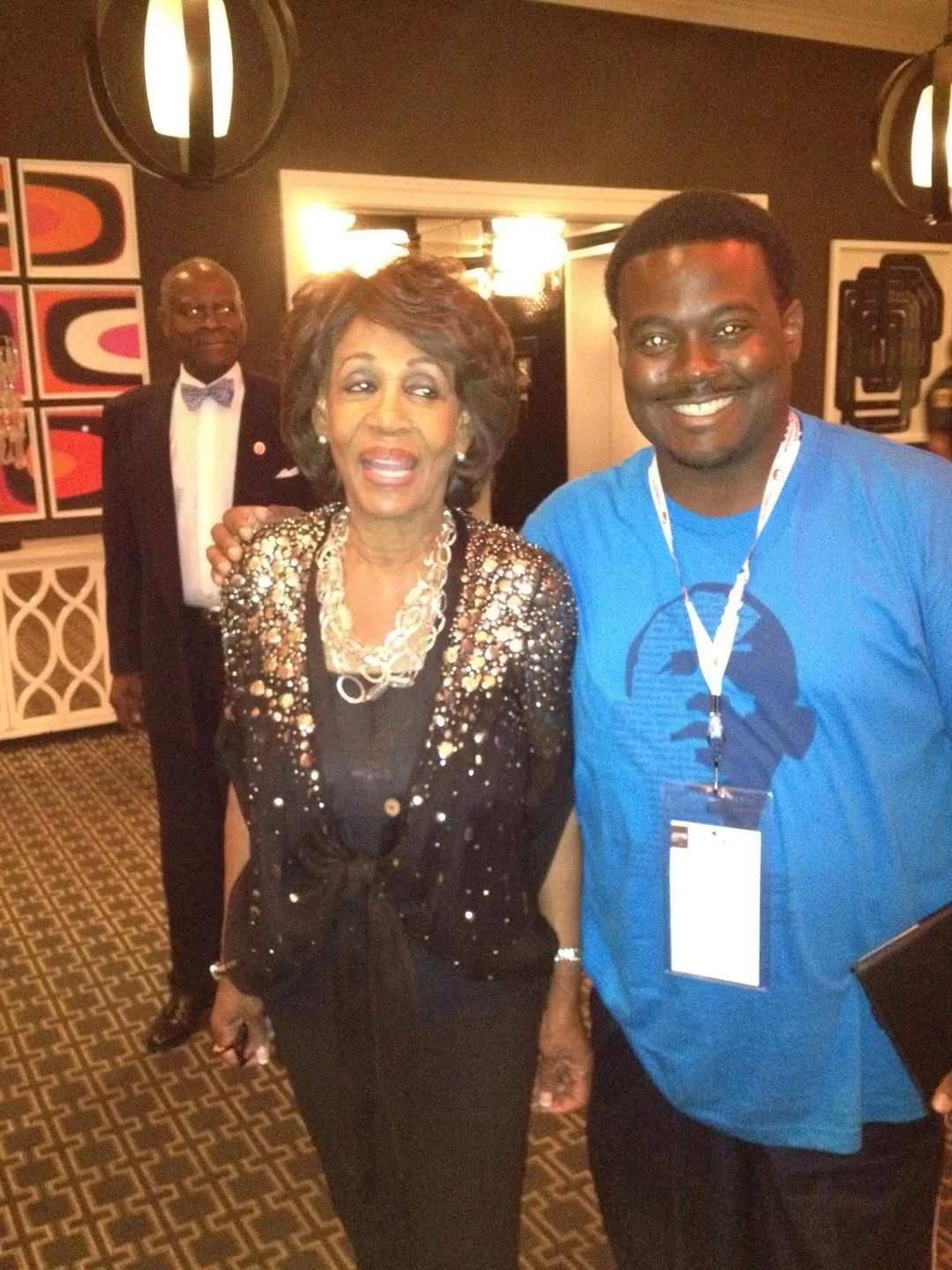 A woman in a black dress with a sequin jacket and layered necklace standing next to a young man in a blue T-shirt with a white lanyard, at an indoor event with a man in a black tuxedo in the background.