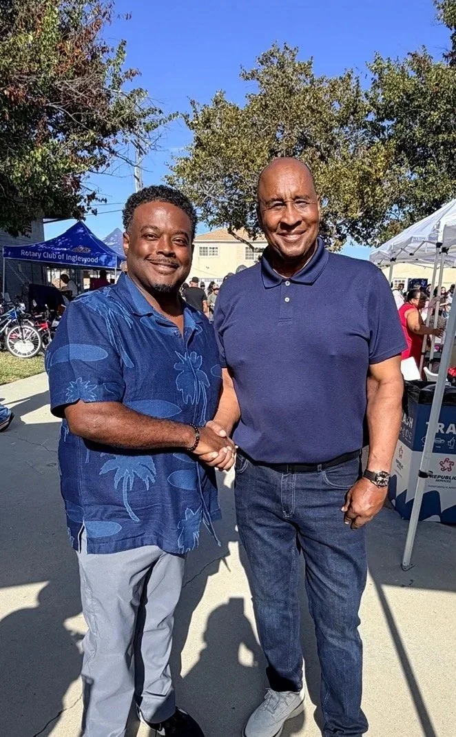 Two men shaking hands outdoors at a community event, smiling, with tents and other people in the background, under clear blue sky.