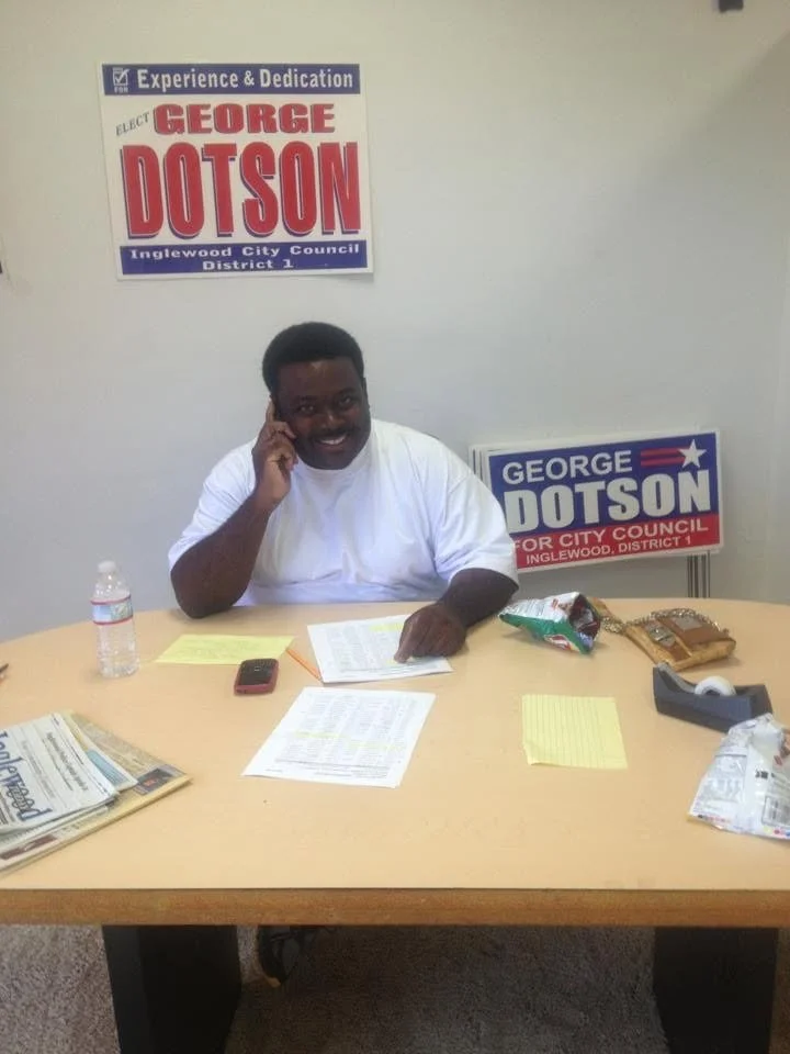 A man sitting at a table with campaign materials, a water bottle, snacks, and papers, smiling and touching his ear, with campaign signs for George Dotson on the wall behind him.