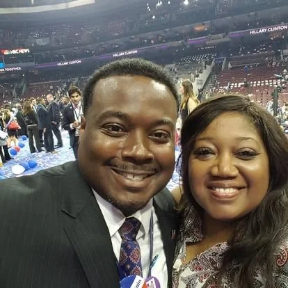 A man and woman smiling for a selfie at a large indoor event with a crowd and stage in the background.