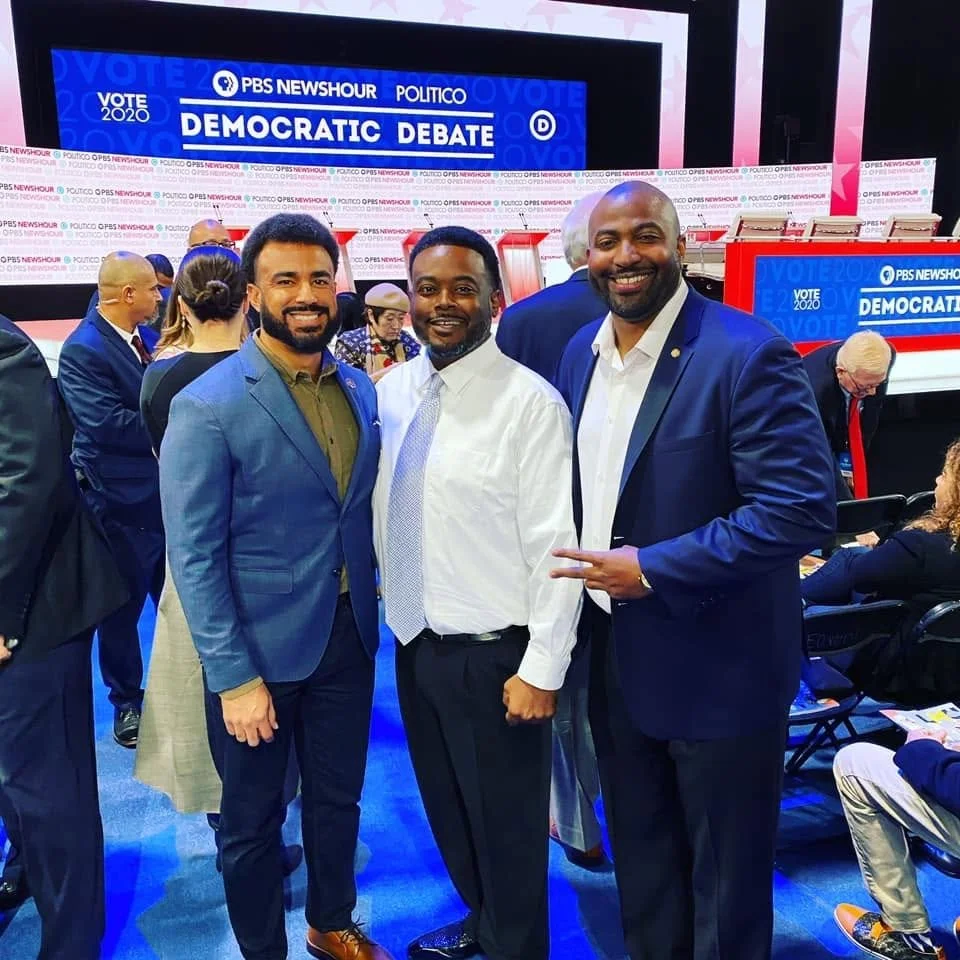 Three men in suits standing together at a Democratic debate event with a large screen in the background showing the words "PBS Newshour" and "Politico" during the 2020 election cycle.