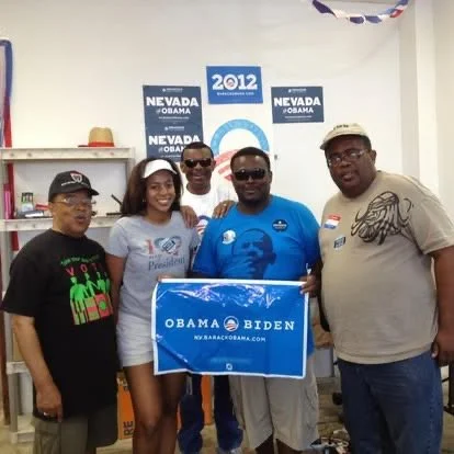 Group of five people standing together indoors holding a blue Obama-Biden campaign sign, with Nevada and 2012 campaign posters in the background.