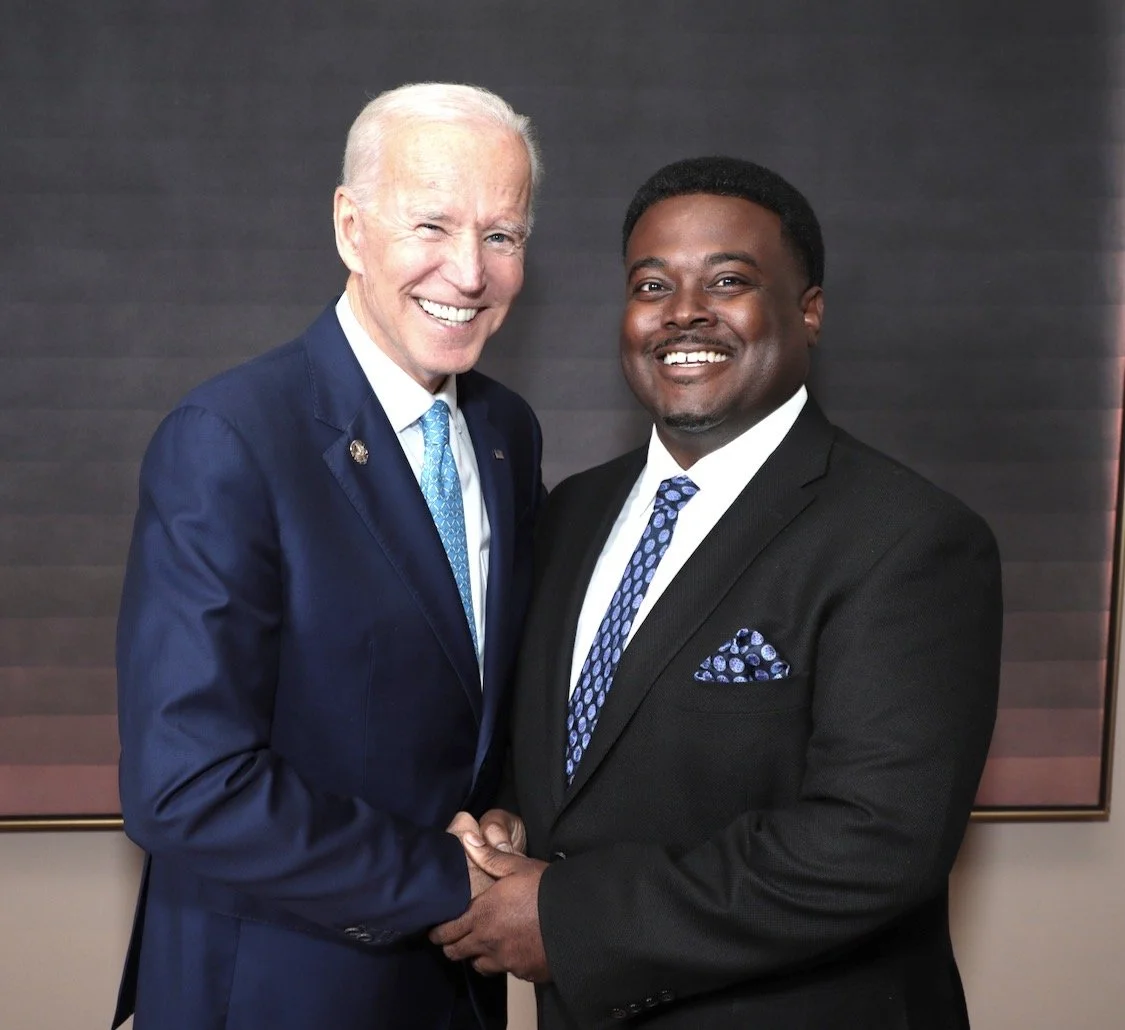 Two men in suits smiling and shaking hands, standing in front of a dark gray background.