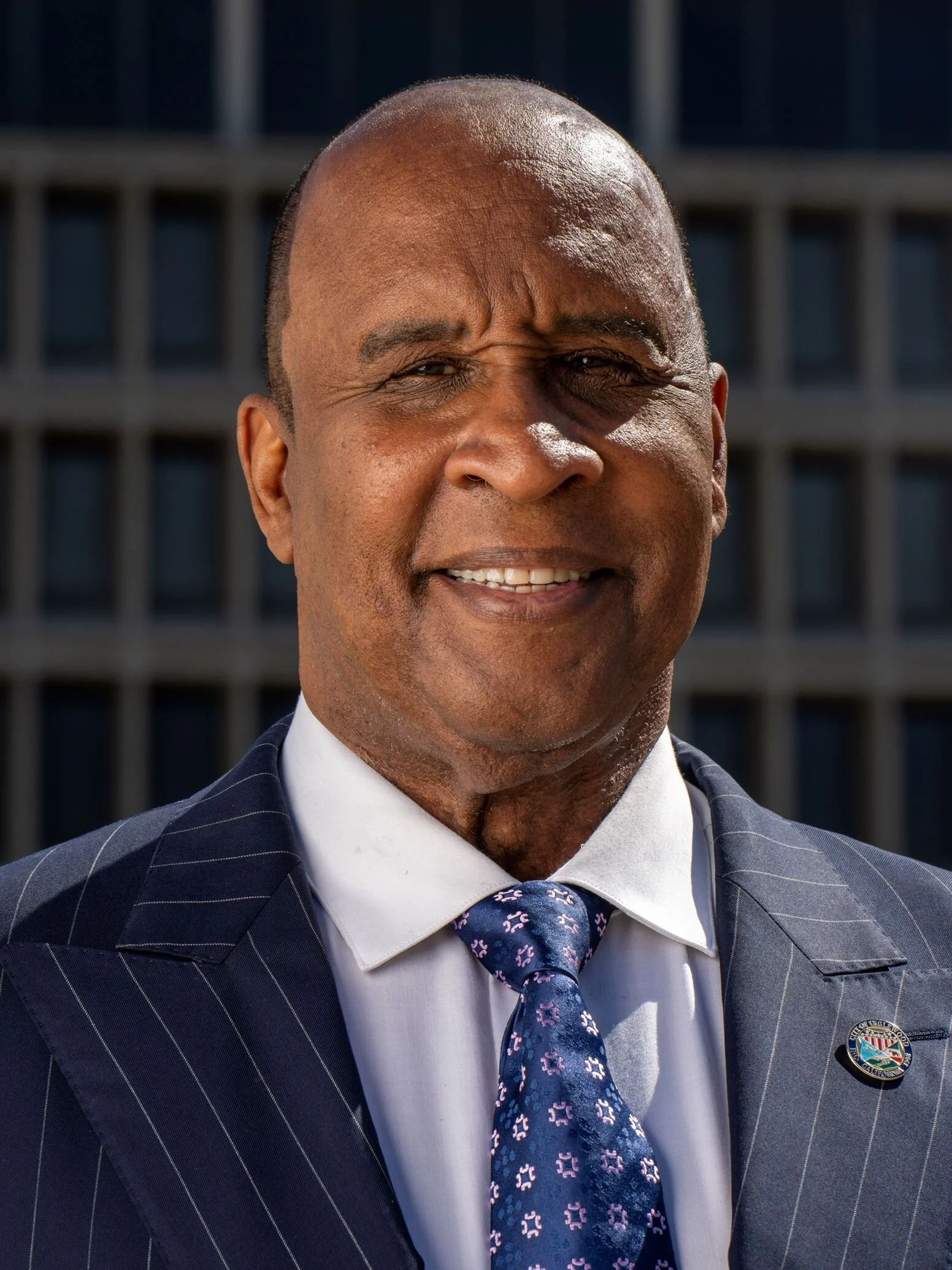 Close-up of a smiling man wearing a pinstripe suit, white shirt, and tie, with a city building background.