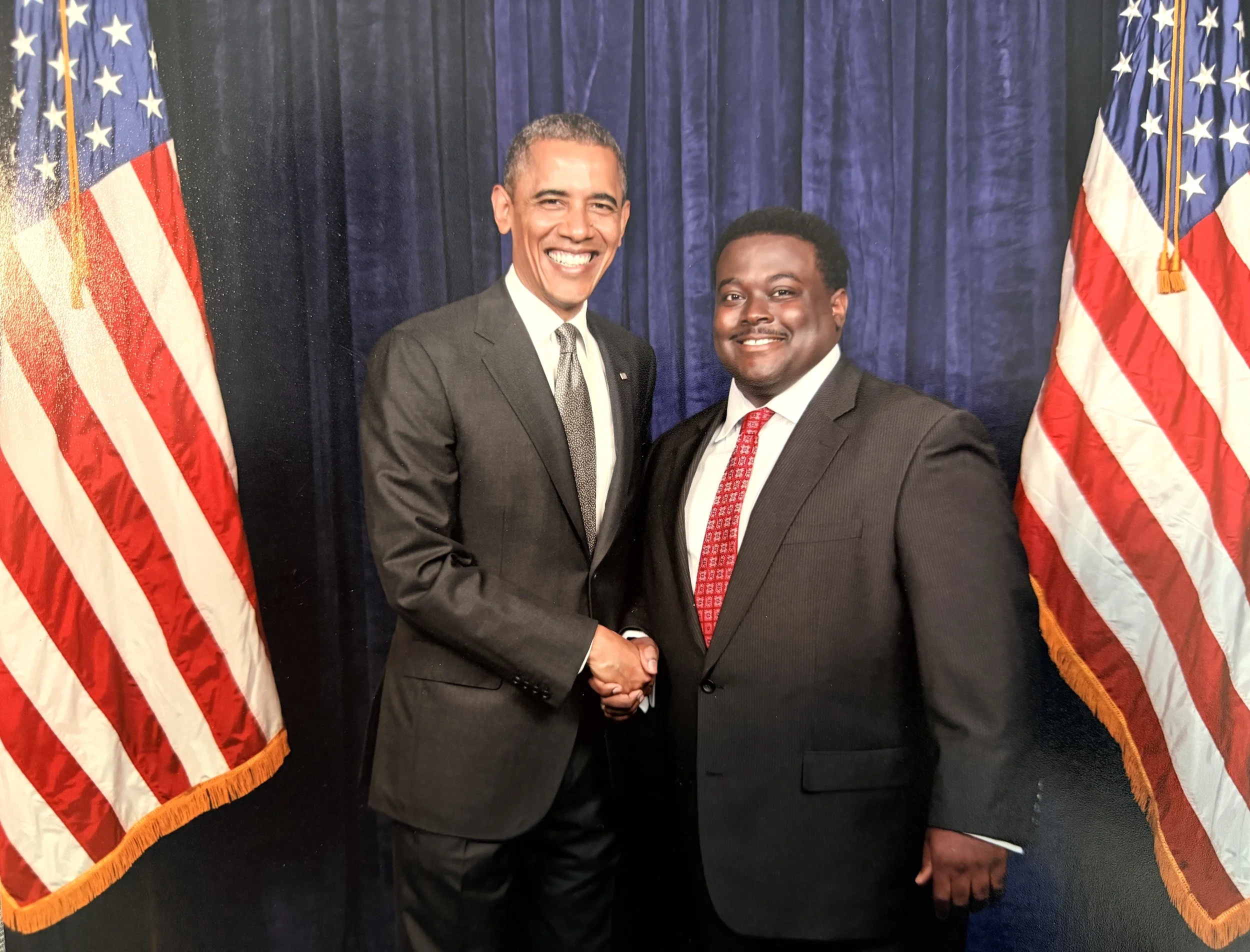 Two men in suits shaking hands, smiling, standing in front of blue curtains and American flags.