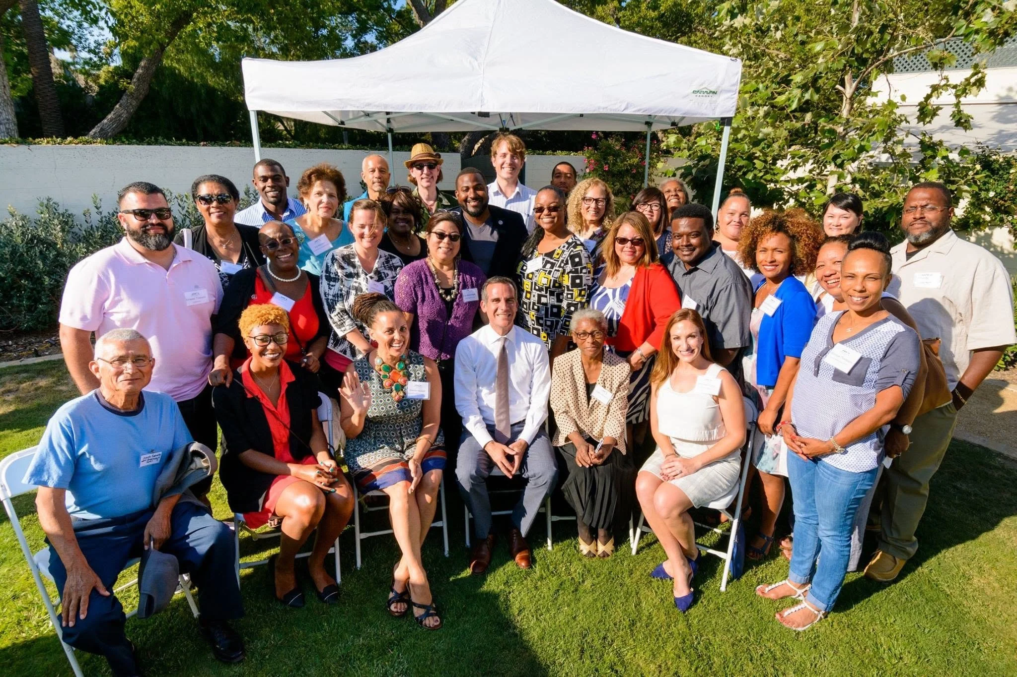 A diverse group of people gathered outdoors under a white canopy, smiling for a group photo on a sunny day with green trees and bushes in the background.