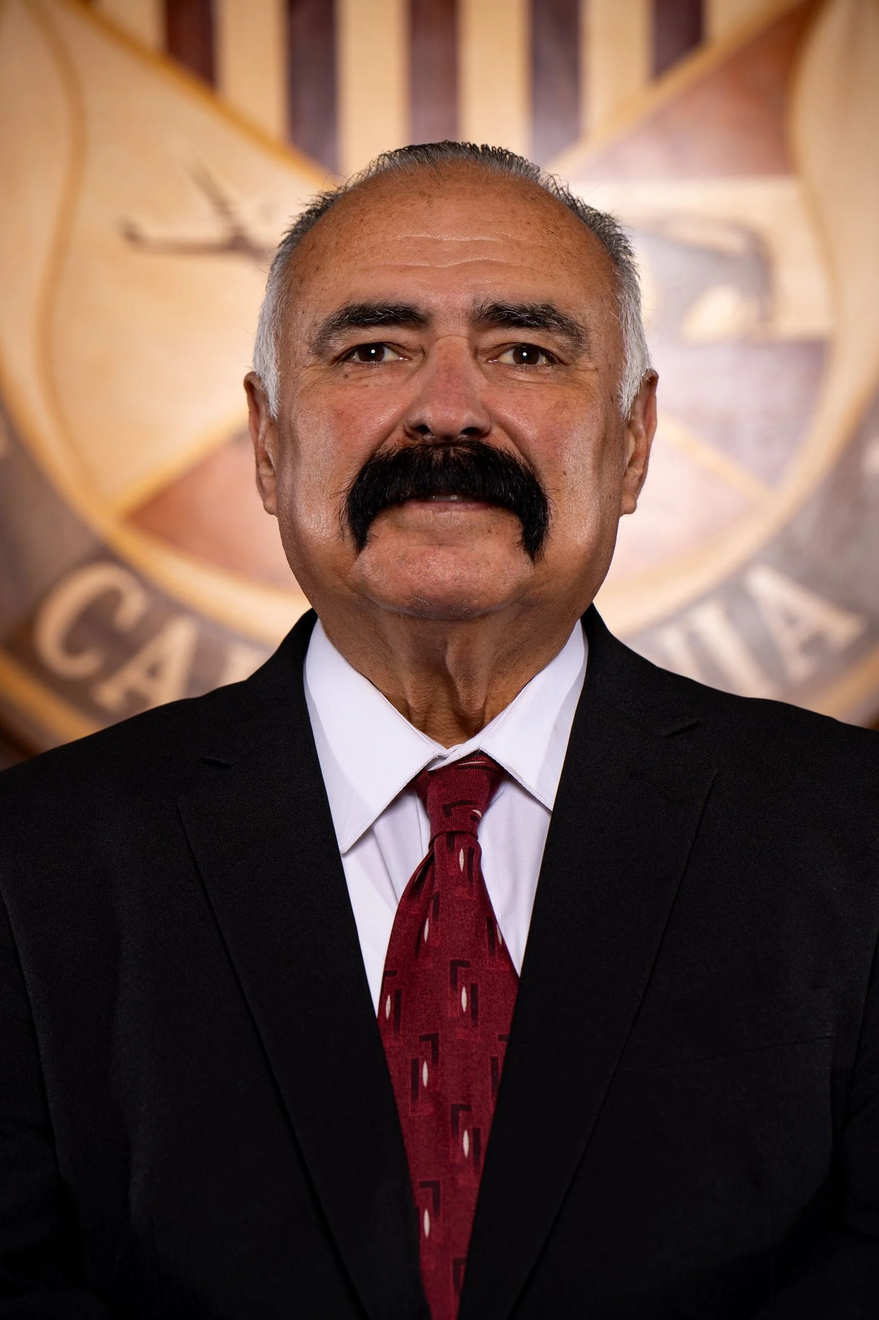 A man with a large black mustache and graying hair wearing a black suit, white shirt, and red tie, standing in front of a wall with a large emblem or seal.