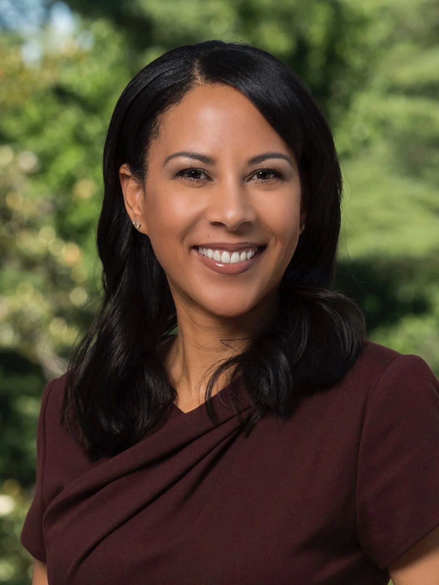 Close-up of a woman with black hair, smiling, wearing a maroon top and earrings, outdoors with a blurred green background.
