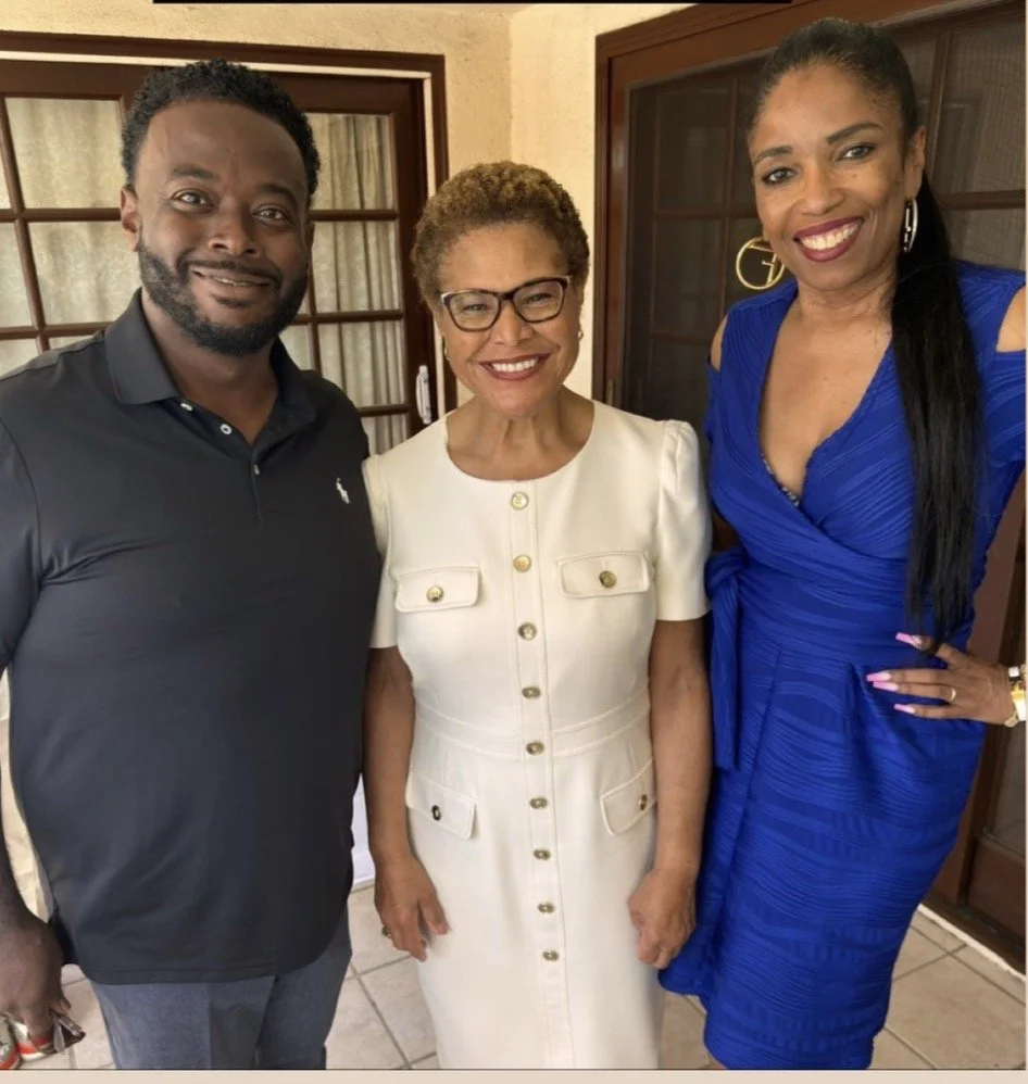 Three people standing together, smiling, inside a house with wooden framed glass doors in the background. The person on the left is a man with short curly hair, wearing a black polo shirt. The person in the middle is an elderly woman with glasses, wearing a cream-colored dress with gold buttons. The person on the right is a woman with long dark hair, wearing a vibrant blue dress with cut-out shoulder details.