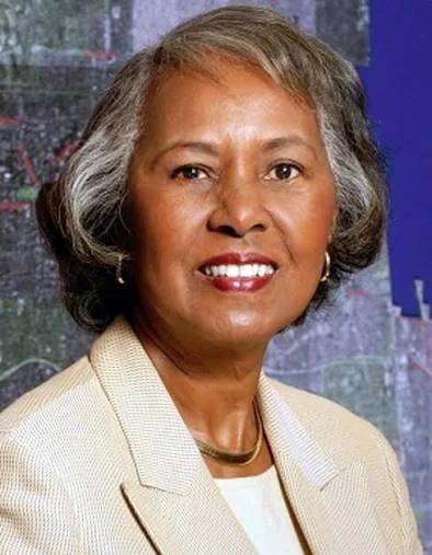 A professional woman with short gray hair, wearing a cream-colored blazer and jewelry, smiling at the camera.