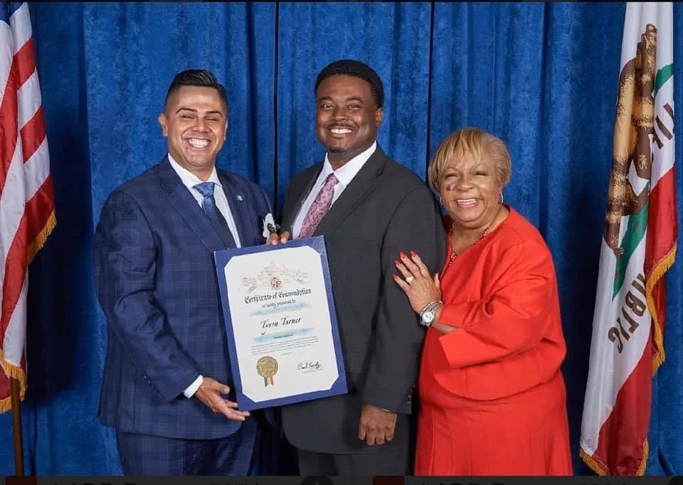 Three people, two men and one woman, standing in front of blue curtains with flags on either side. The man on the left is holding a certificate, and everyone is smiling.