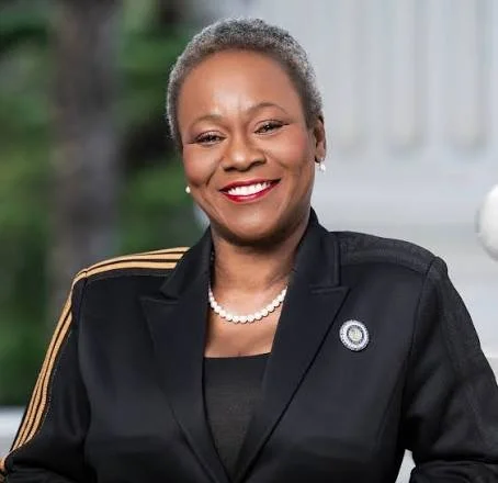 Portrait of a smiling woman in a black blazer with gold stripes on the shoulders, wearing pearl earrings and a pearl necklace, standing outdoors with a blurred background.