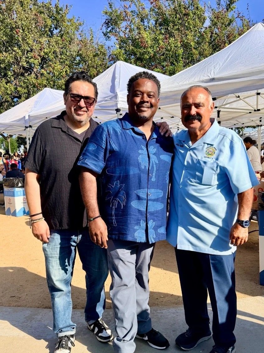 Three men standing outdoors at a daytime event with tents in the background. The man on the left has dark hair, glasses, and is wearing a black shirt. The man in the middle has short curly hair and is dressed in a blue Hawaiian shirt. The man on the right is wearing a light blue uniform shirt with a badge. All three are smiling and posing for the photo.