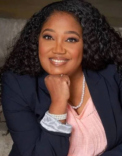 A woman with curly black hair, wearing a navy blazer, pink top, pearl necklace, and bracelet, resting her chin on her hand and smiling.